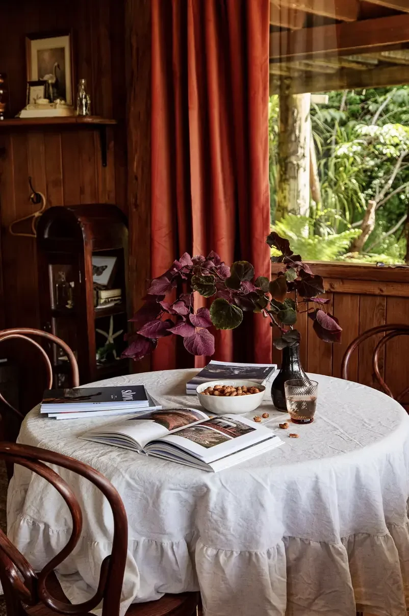 Round dining table with white linen tablecloth and bentwood chairs, terracotta velvet curtains and lush botanical garden beyond, Del Sol Treehouse Rosny Tasmania