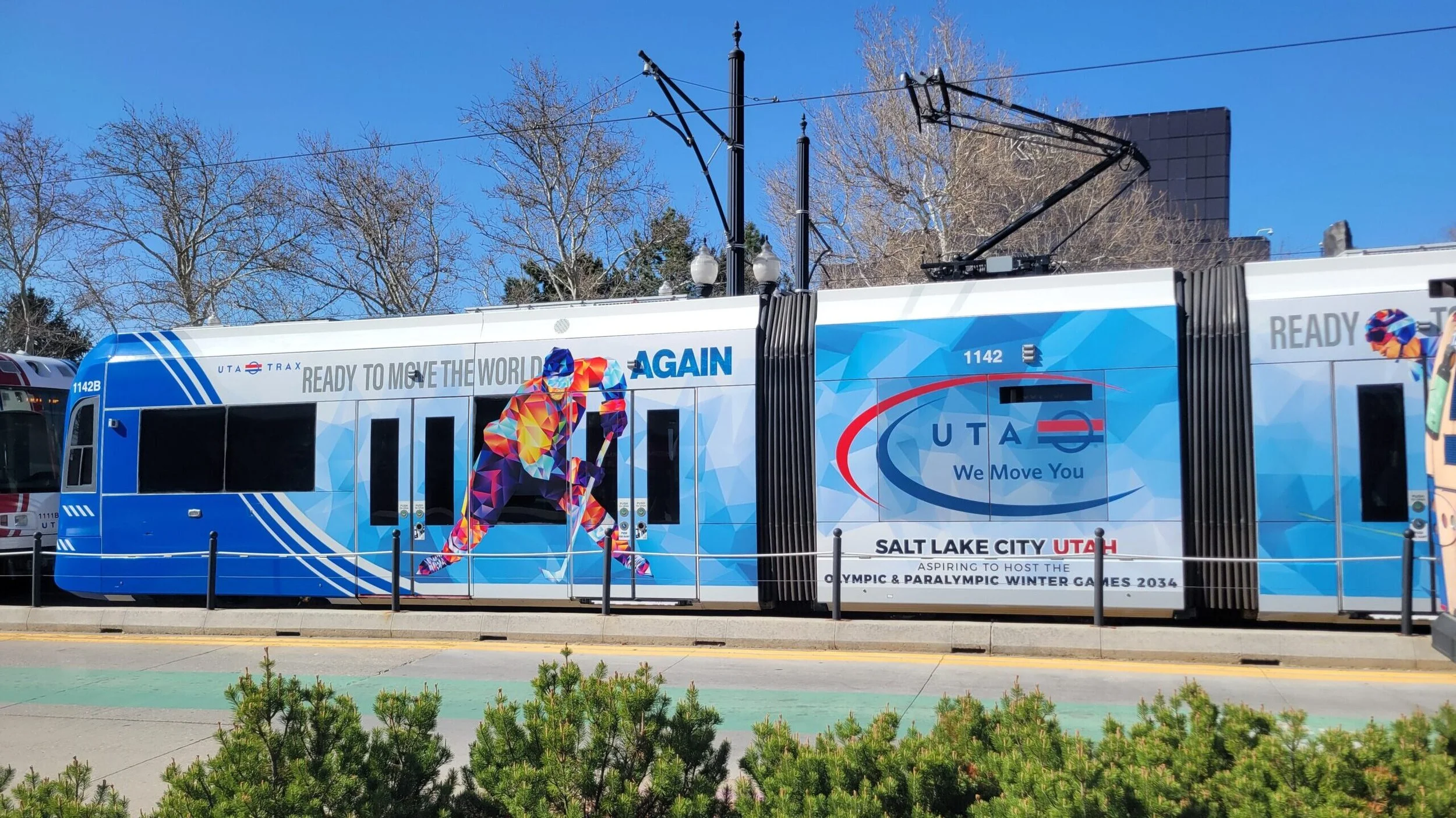 A tram decorated with blue graphics and advertisements, featuring a colorful image of a hockey player and the text "Ready to Move the World Again". The tram is parked outdoors on a sunny day with leafless trees and a blue sky in the background.