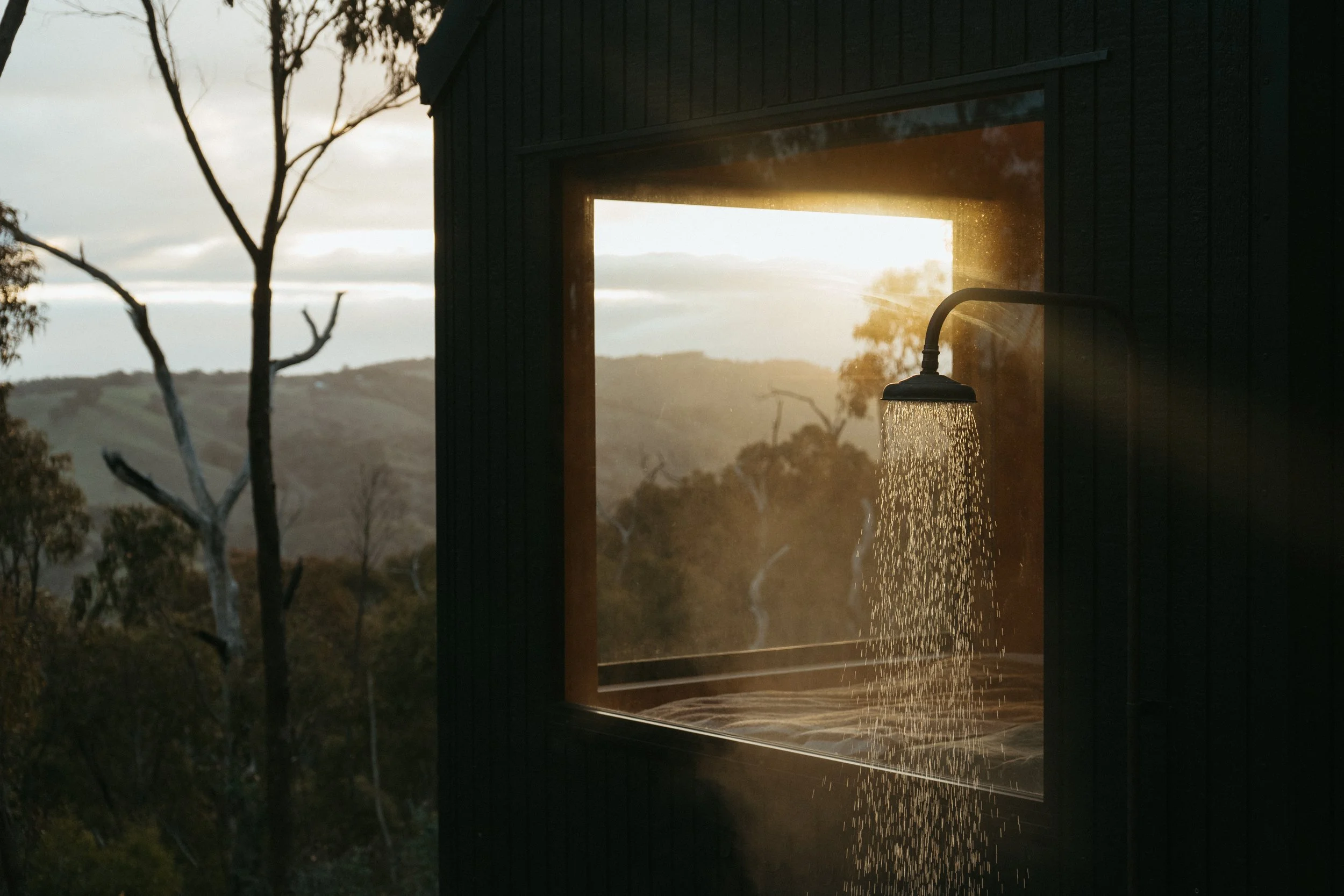 Showerhead with water flowing outside a window during sunset, with trees and hills in the background.