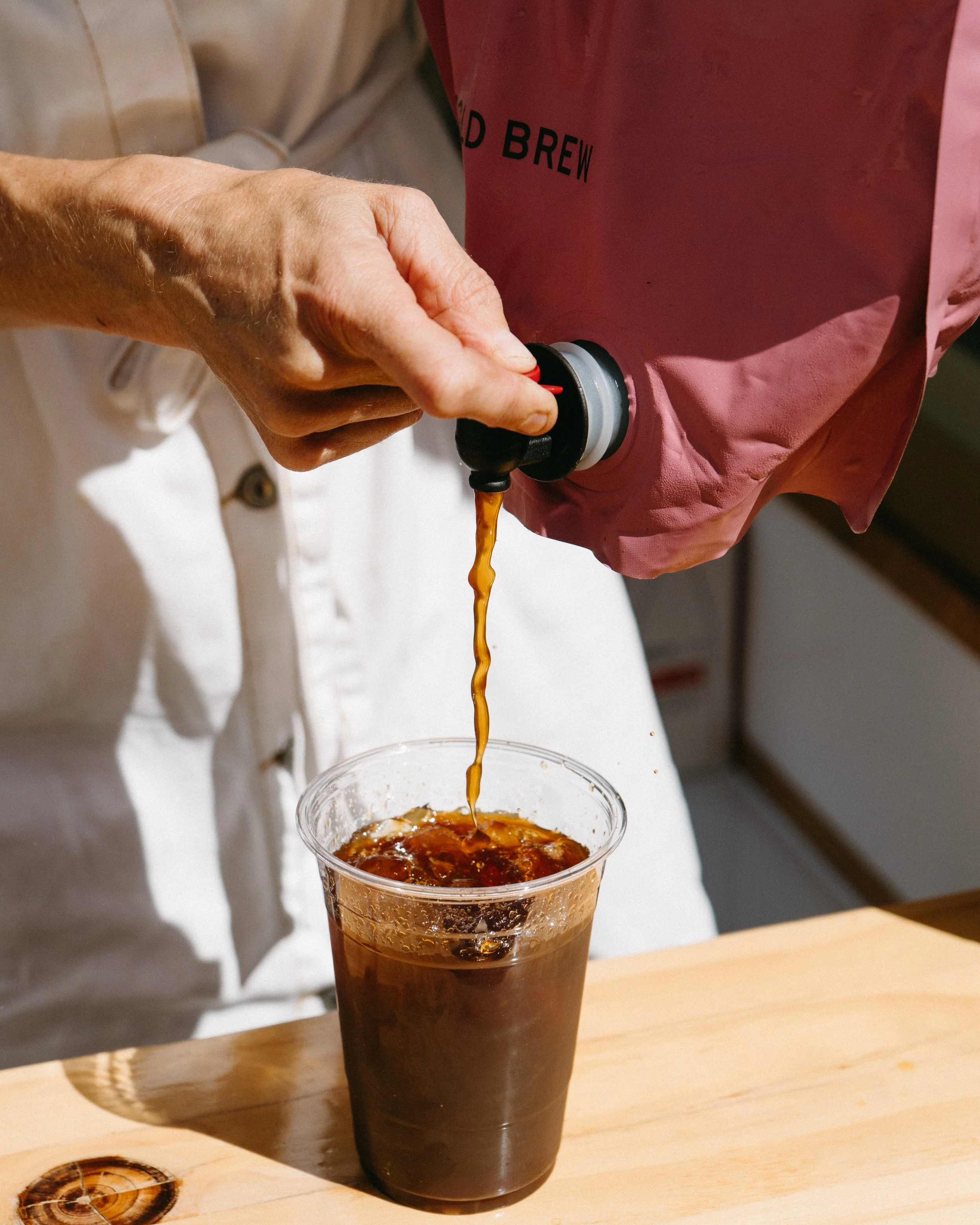 A person pouring cold brew coffee from a pink bag into a clear plastic cup filled with ice on a wooden surface.