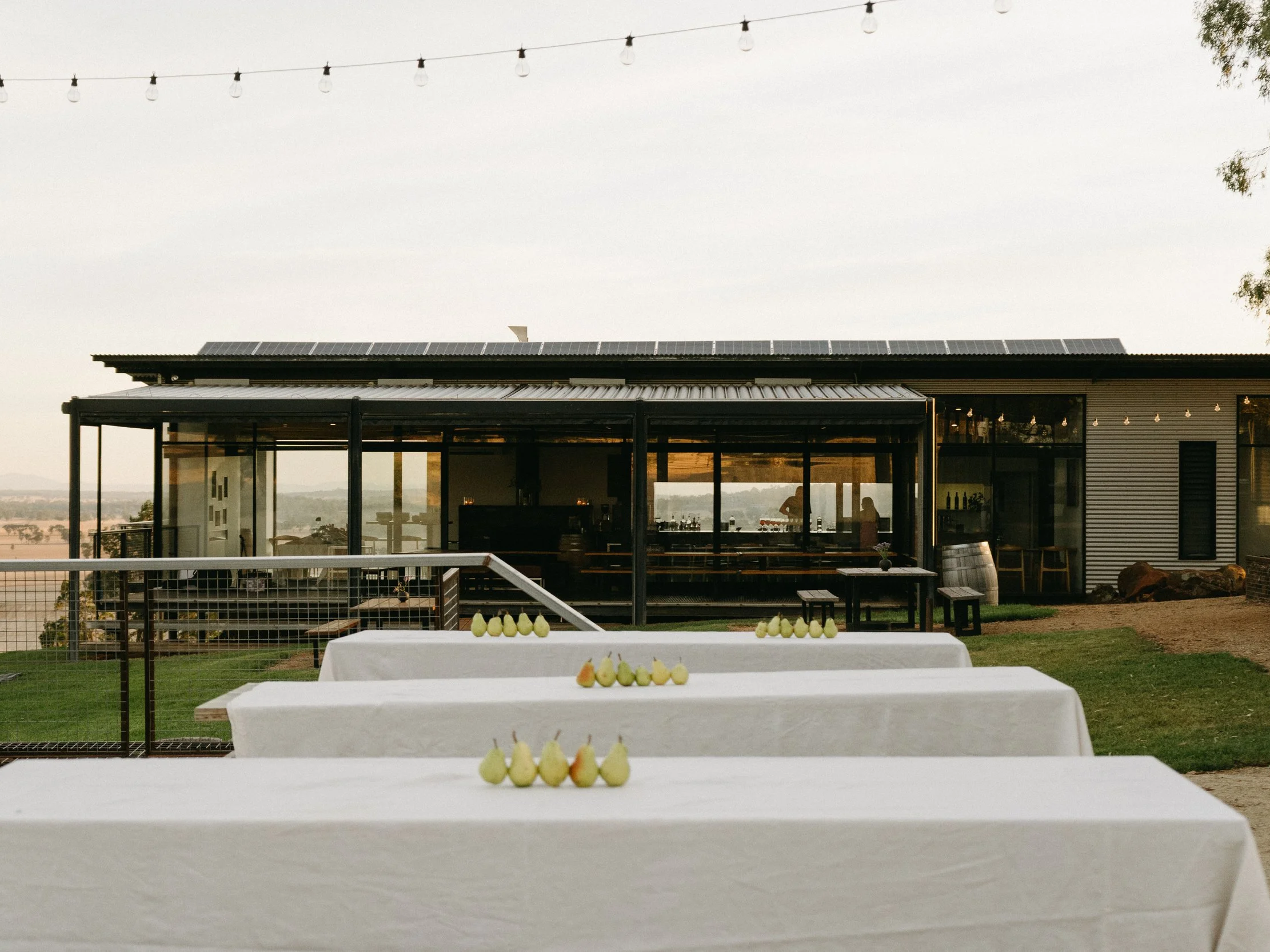 Outdoor event setup with tables covered in white tablecloths and small green pears decorations, near a modern building with large windows and string lights overhead, overlooking a scenic landscape.