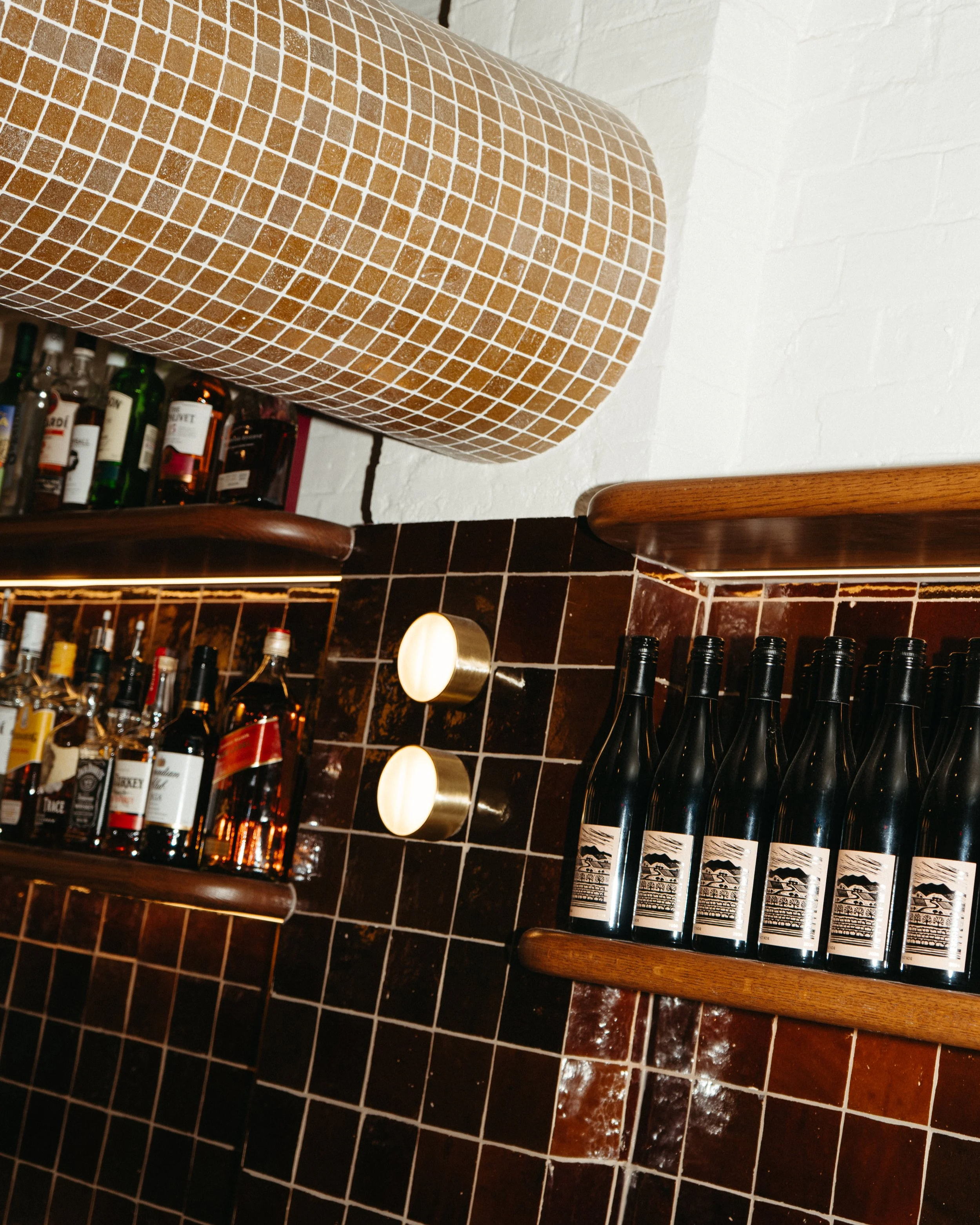 A view of a bar shelf with various liquor bottles, a wall with dark brown tiles, and a wooden shelf with black bottles with white labels, and a wall-mounted light fixture.