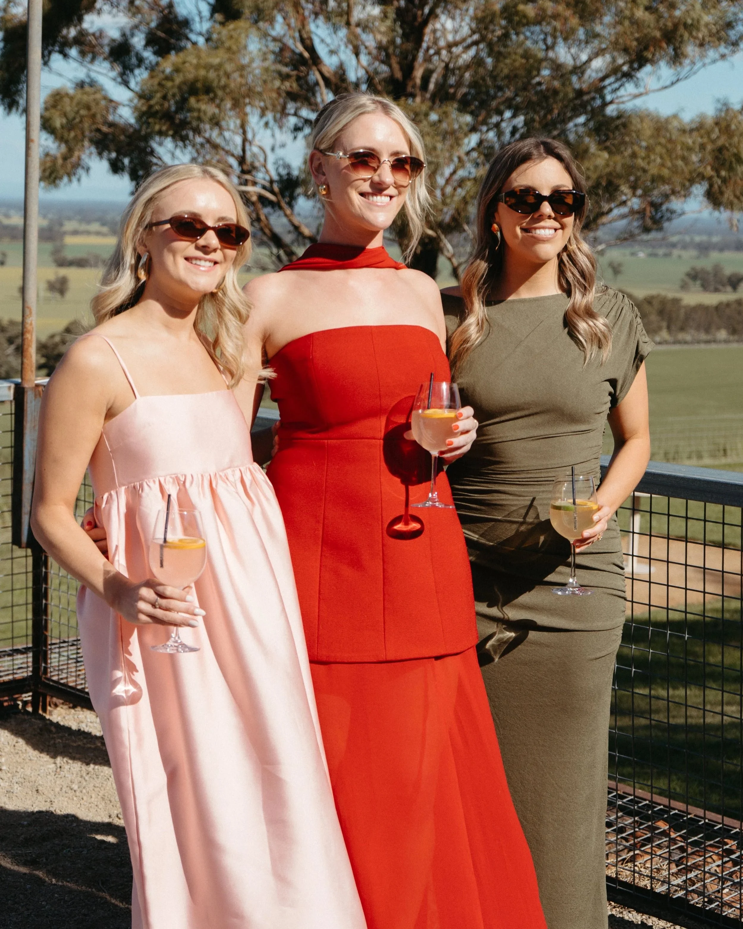 Three women standing outdoors, dressed in elegant dresses, holding glasses of drinks with lemon slices, smiling at the camera on a sunny day.