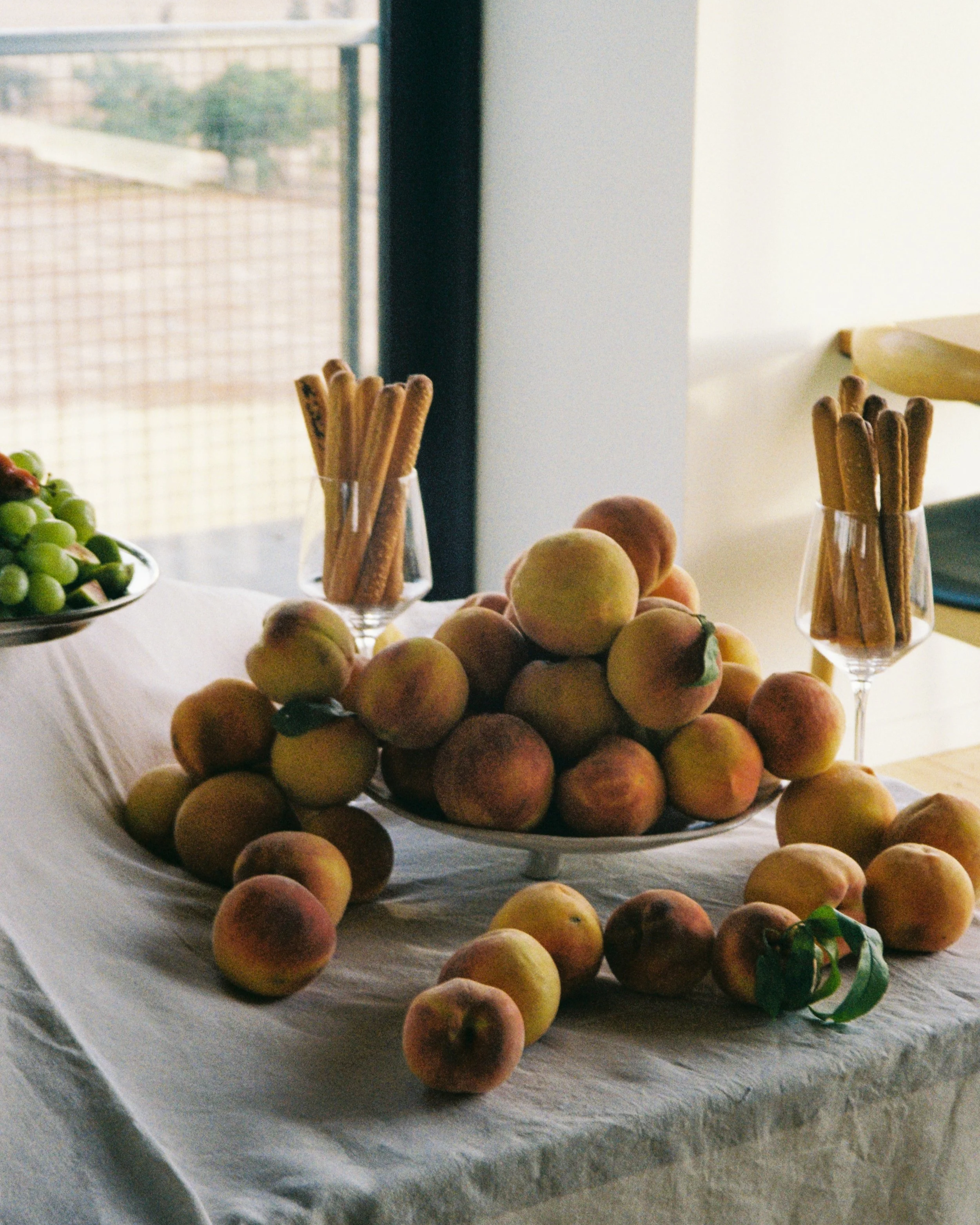 A table with a large bowl of peaches, two glass holders with breadsticks, and a small plate of green grapes in the background, near a window.