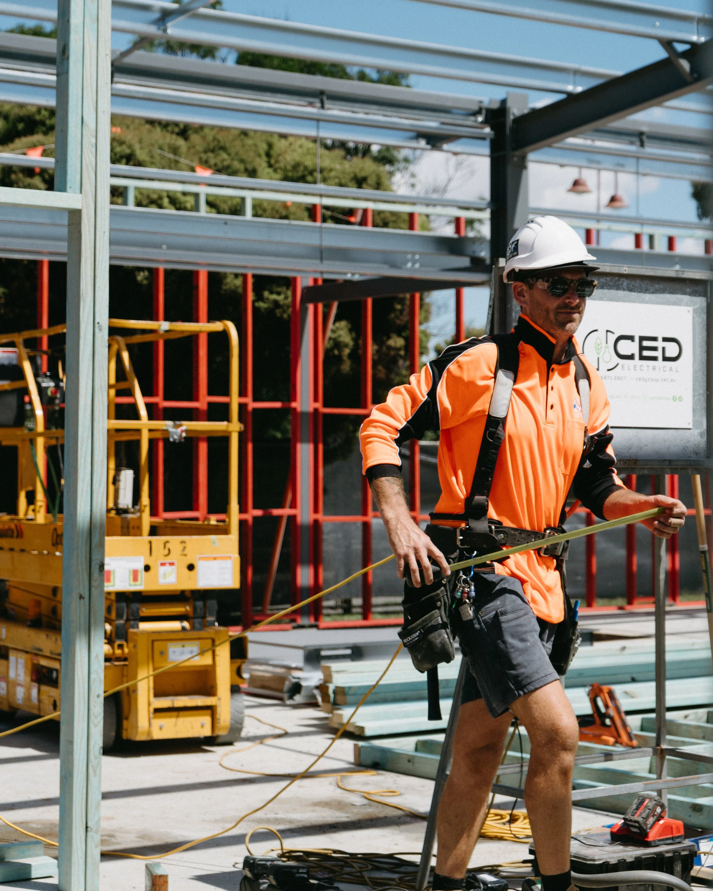 Construction worker in safety gear measuring at construction site with metal framework and equipment.