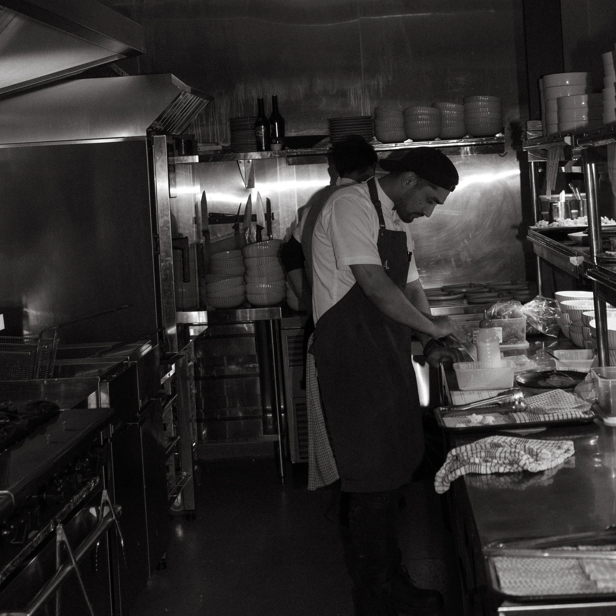 A chef in a professional kitchen preparing food, wearing a black apron and a bandana, with various kitchen utensils, bowls, and ingredients around him.