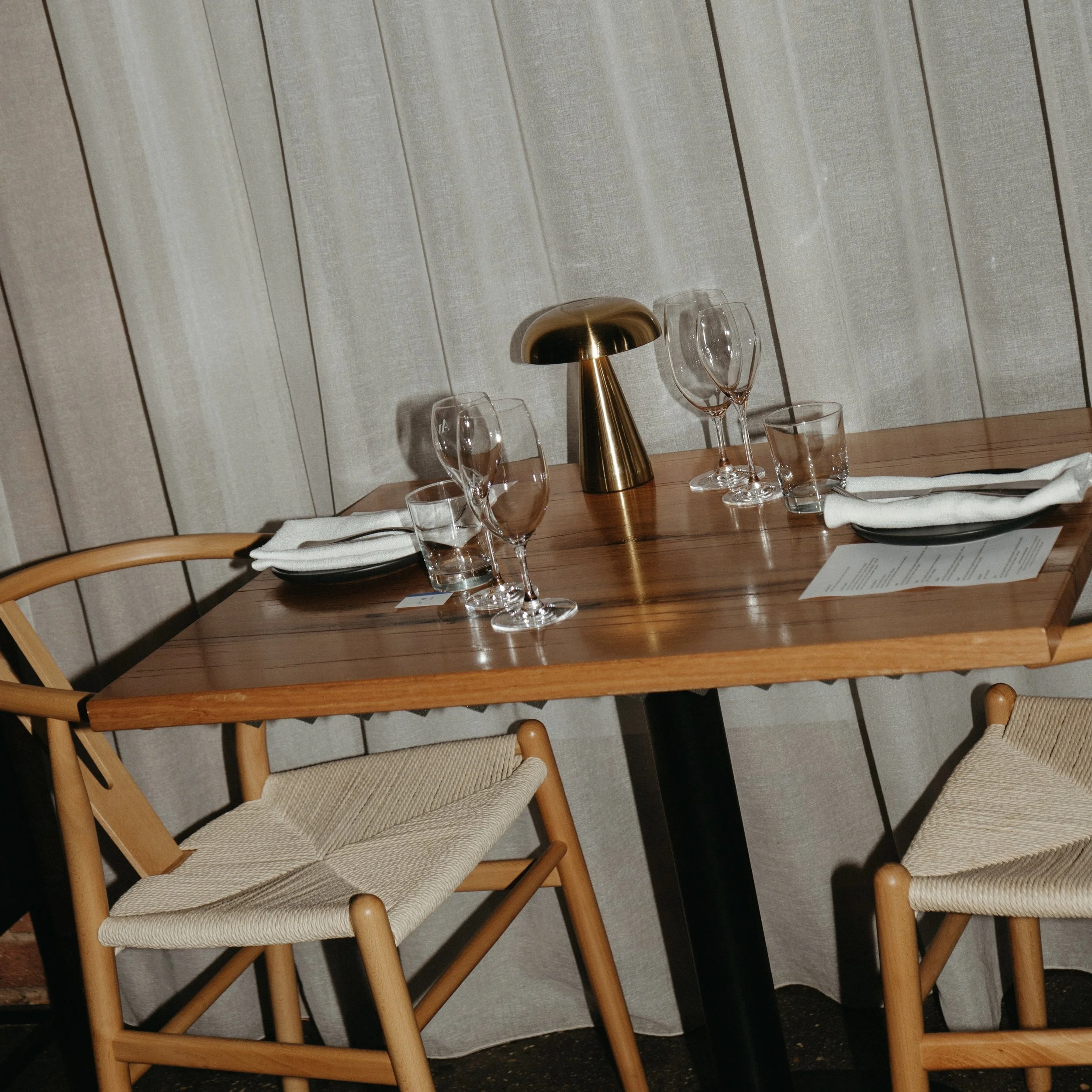 A restaurant table set with two place settings, wine glasses, water glasses, white napkins, and a gold-colored modern table lamp on a wooden table with two chairs.
