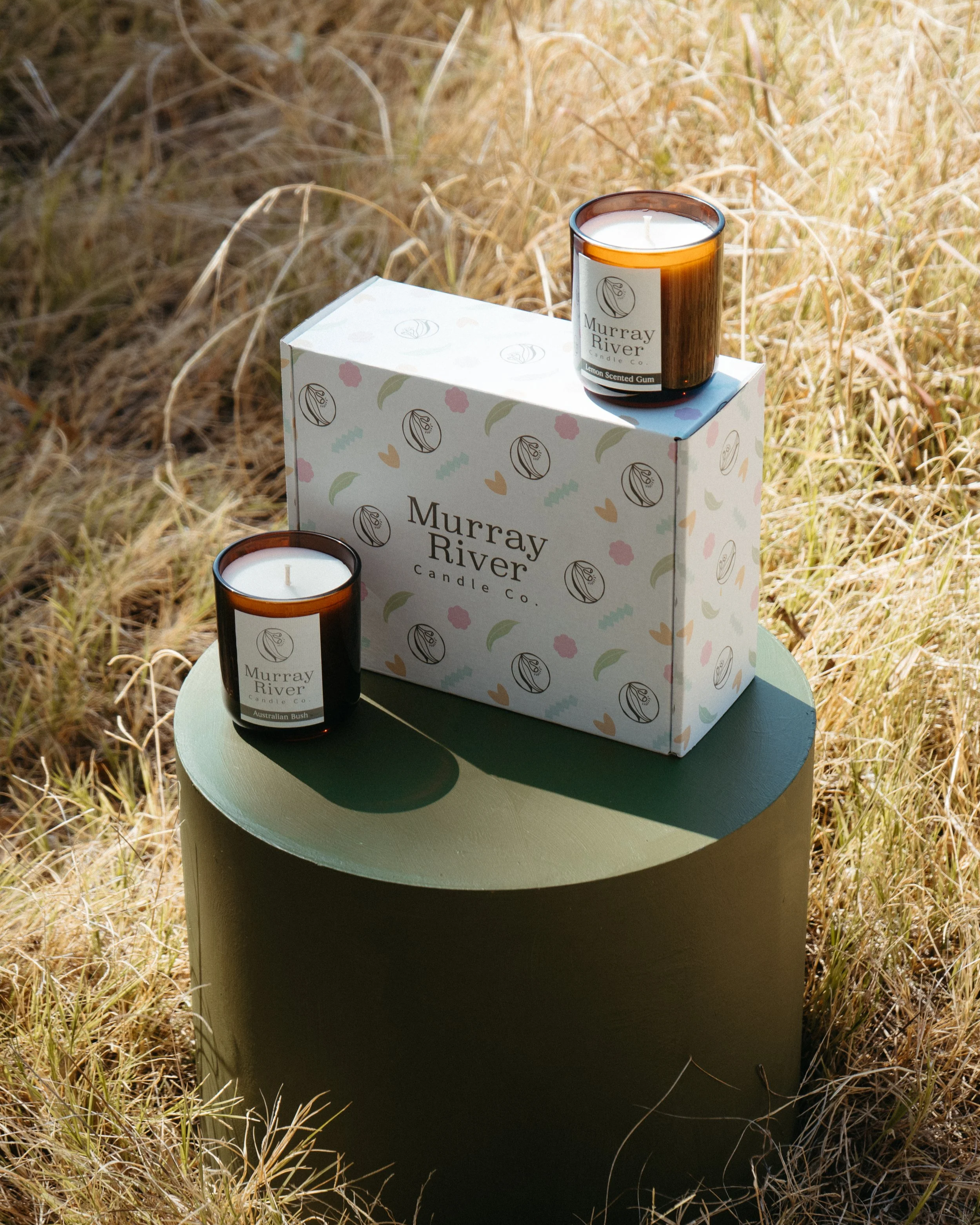 Two scented candles labeled 'Murray River Candle Co.' on a green cylindrical pedestal with a decorative box behind them, set in a grassy outdoor area.