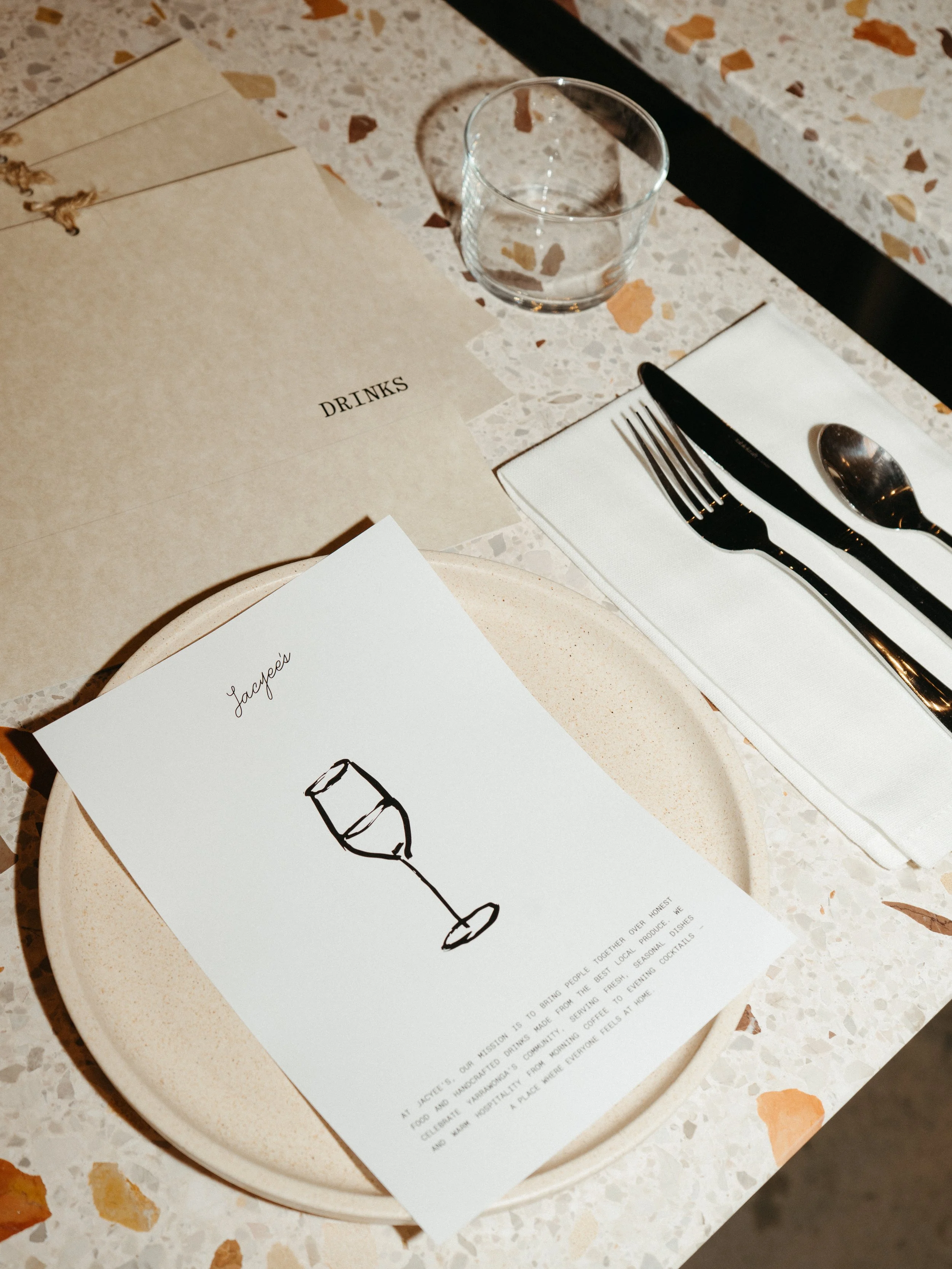 Table setting with a plate, a menu, a glass of water, and cutlery on a terrazzo table.