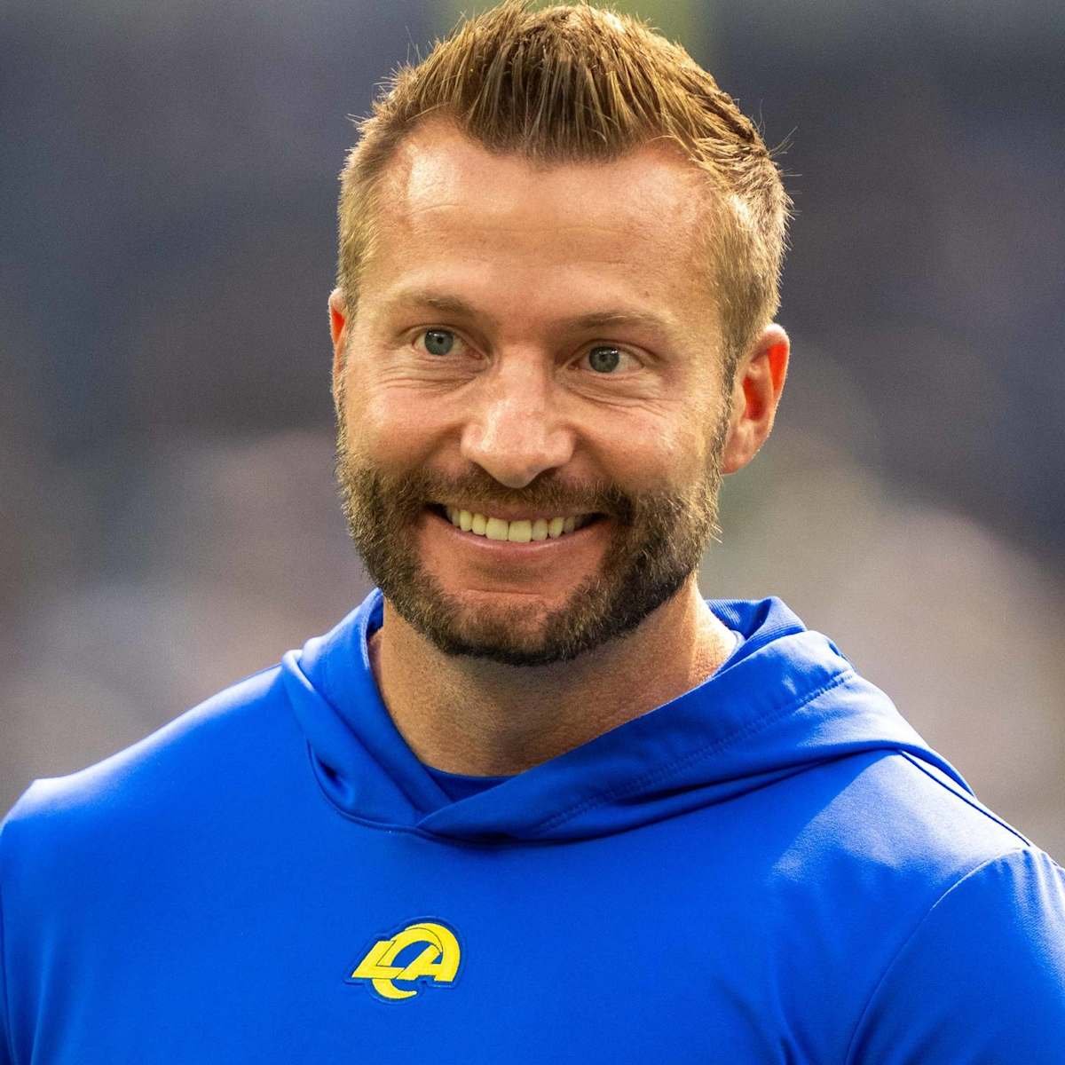 A man with short brown hair, a beard, and blue eyes smiling, wearing a blue sports hoodie with a yellow logo, in an indoor sports setting.