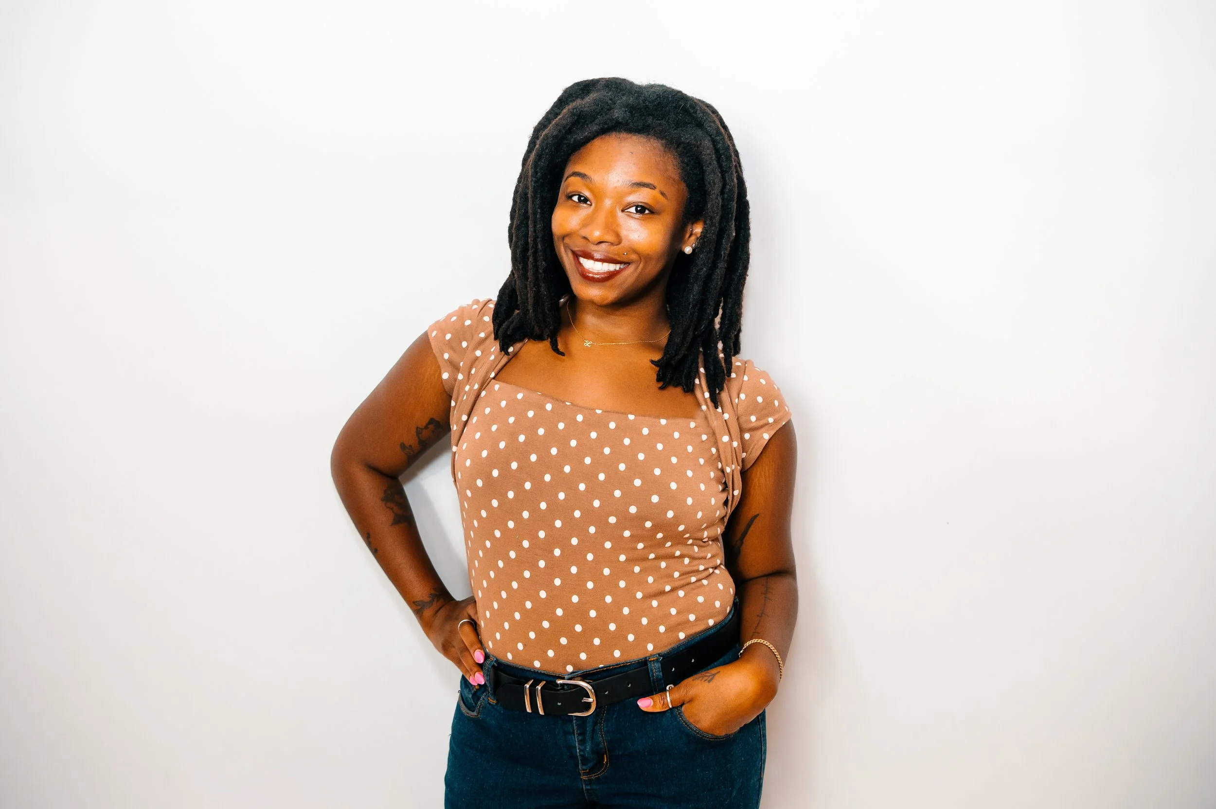 A woman with black dreadlocks, wearing a brown polka dot top and blue jeans, smiling, standing against a plain white background.