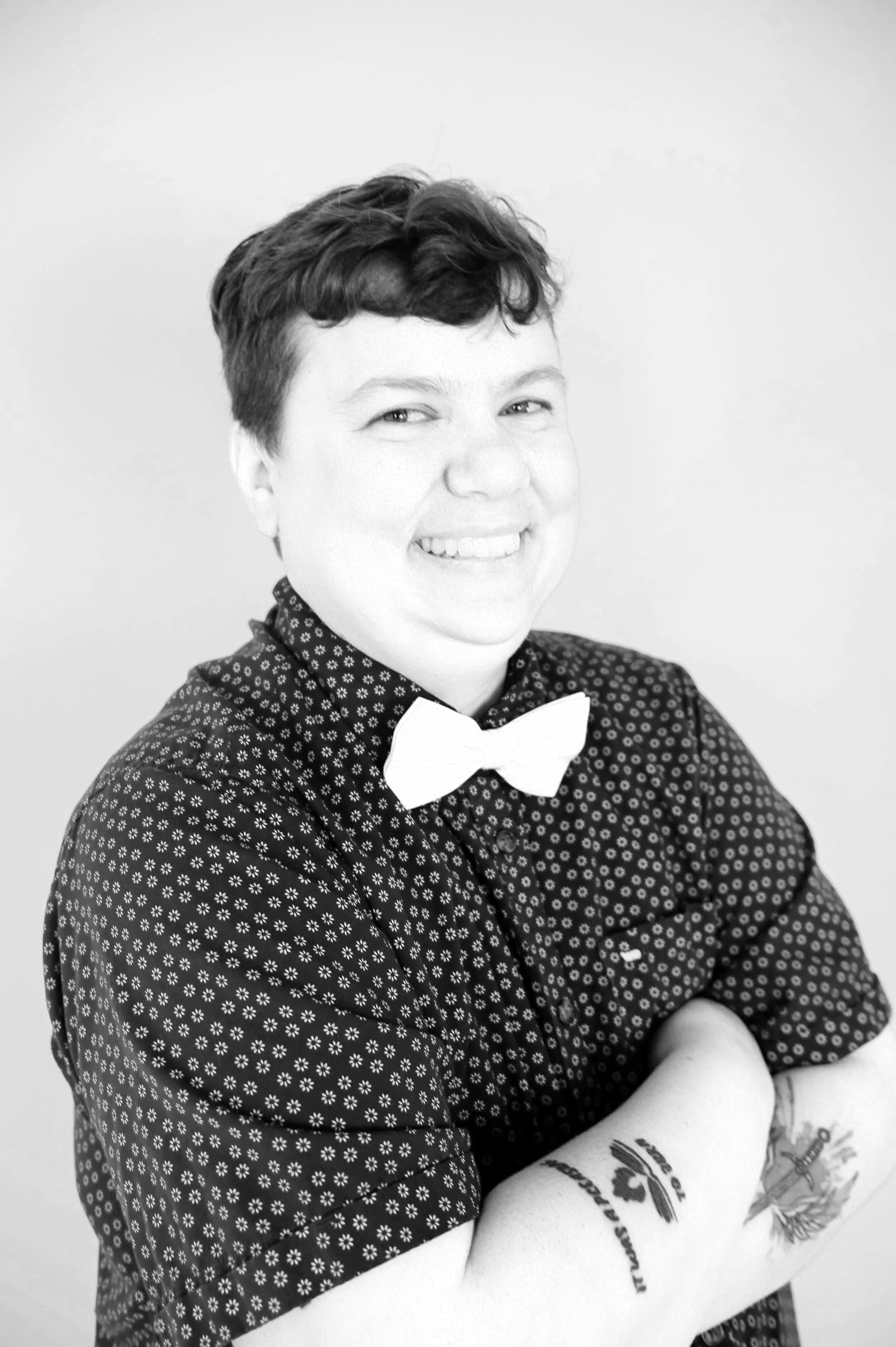Black and white portrait of a smiling person with short curly hair, wearing a patterned button-up shirt, standing against a plain wall.