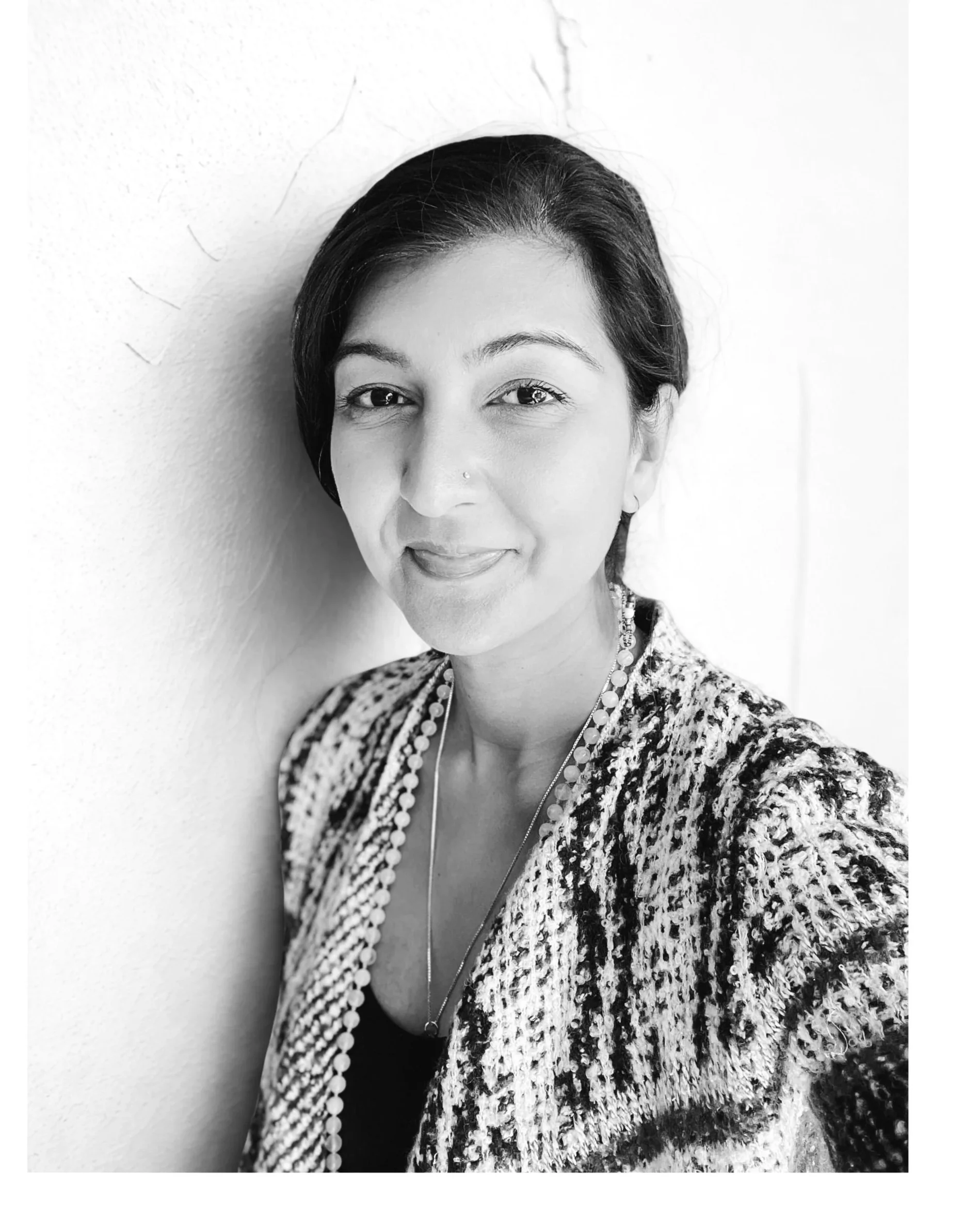 Black and white portrait of a woman with short dark hair, wearing a patterned jacket and layered necklaces, standing against a plain white wall.