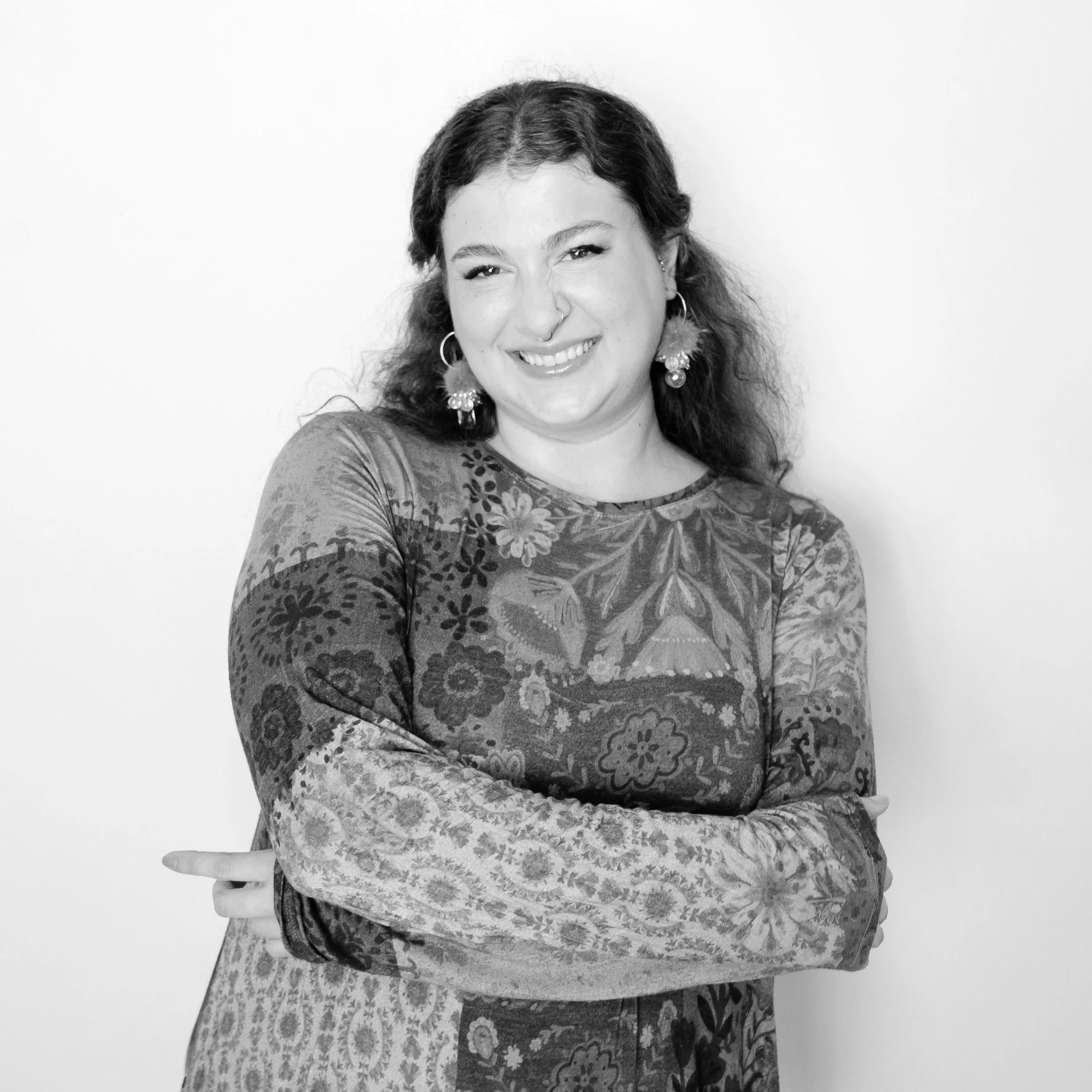 Black and white portrait of a smiling woman with curly hair, wearing floral earrings and a patterned long-sleeve shirt, standing against a plain white wall.