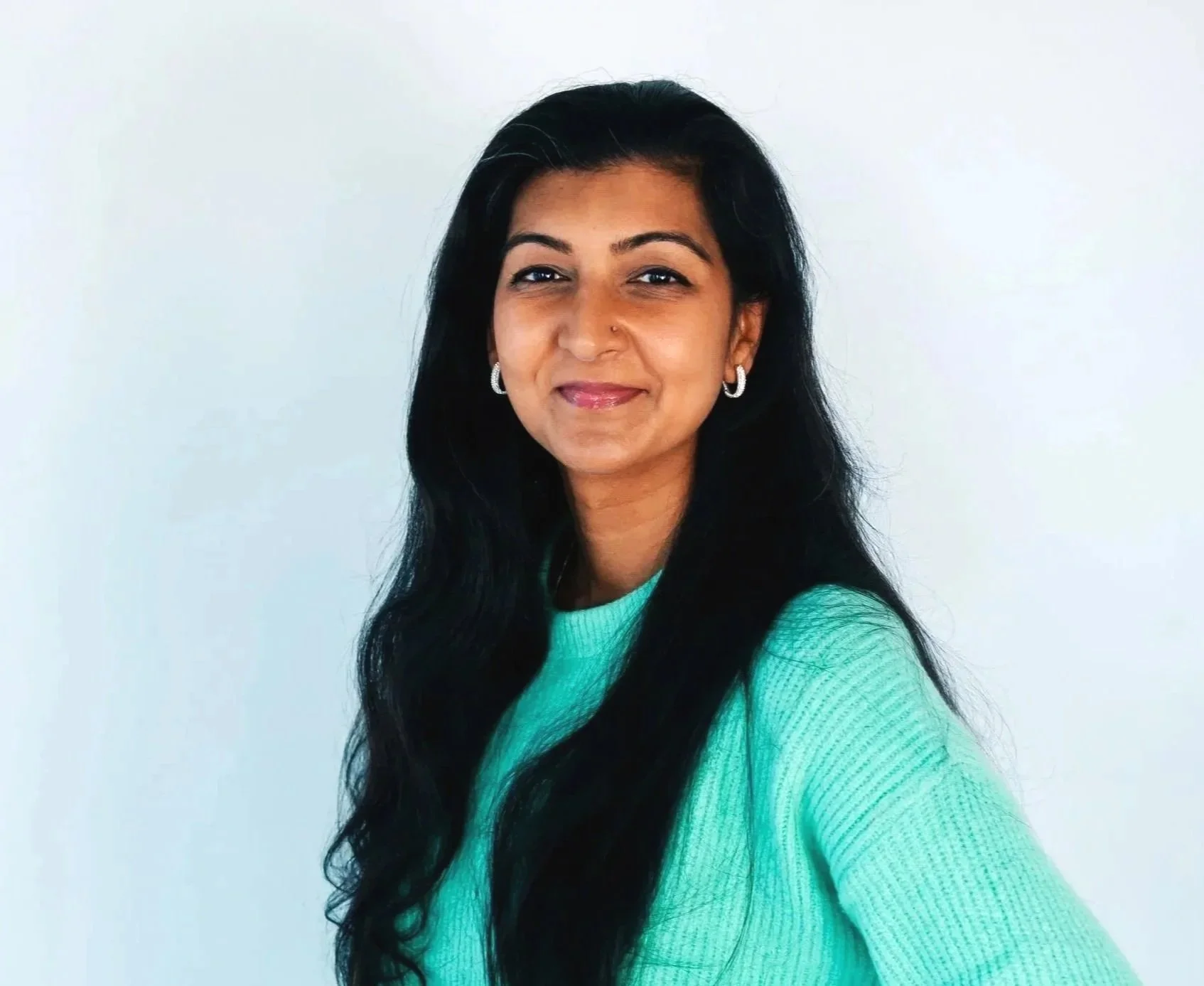 Black and white portrait of a woman with short dark hair, wearing a patterned jacket and layered necklaces, standing against a plain white wall.