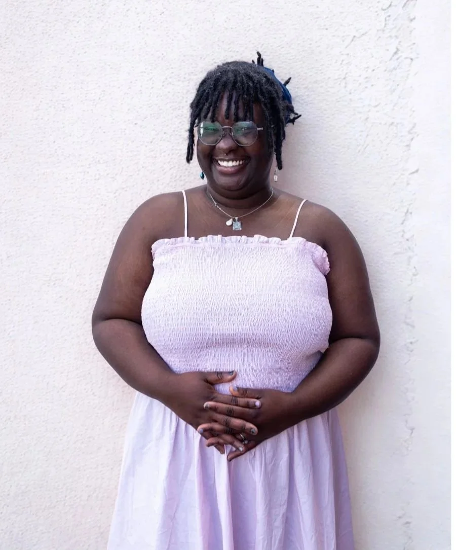 A woman with glasses smiling, dressed in a sleeveless top and skirt, standing against a plain wall.
