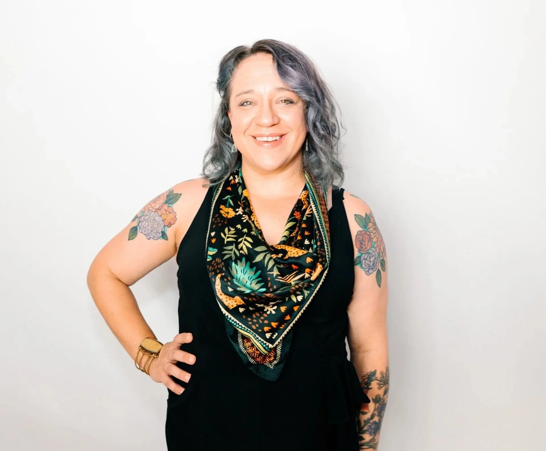 Black and white portrait of a smiling woman with wavy hair, wearing a sleeveless top, a patterned scarf around her neck, a watch, and earrings, with tattoos of flowers on her arms, standing against a plain background.