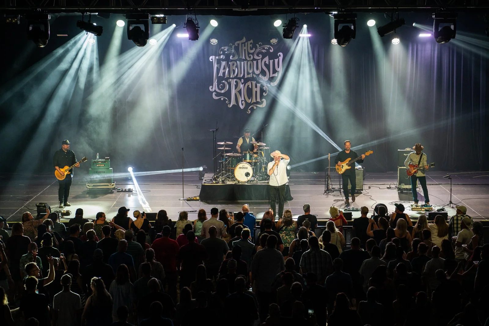 A live performance stage shot with the band performing in front of an audience. The stage has a backdrop with the words 'The Fabulously Rich' and the band includes a drummer, three guitarists, and a lead singer.
