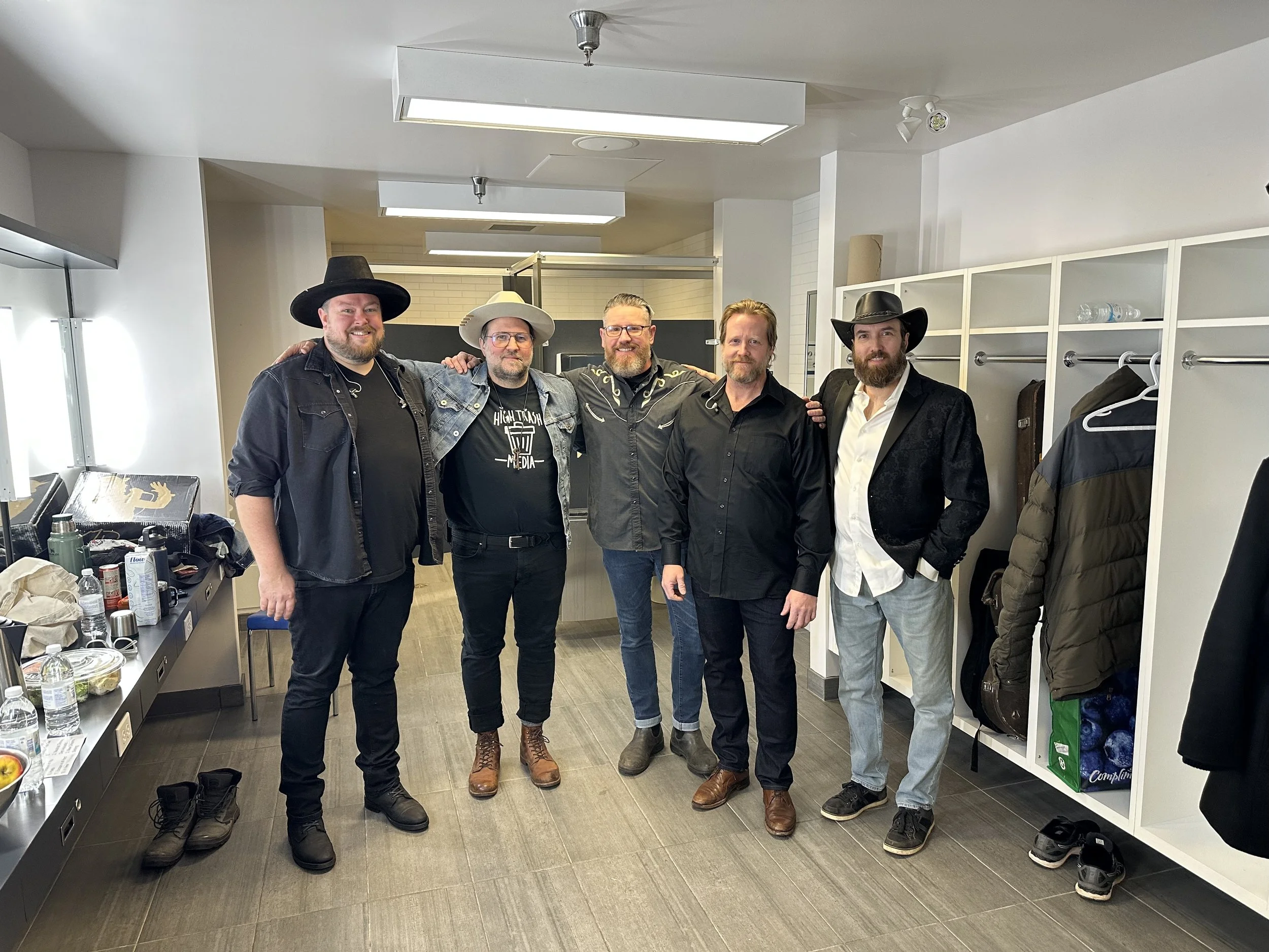 The band backstage standing inside a locker room, some wearing cowboy hats, posing for a photo. They are smiling and have their arms around each other.