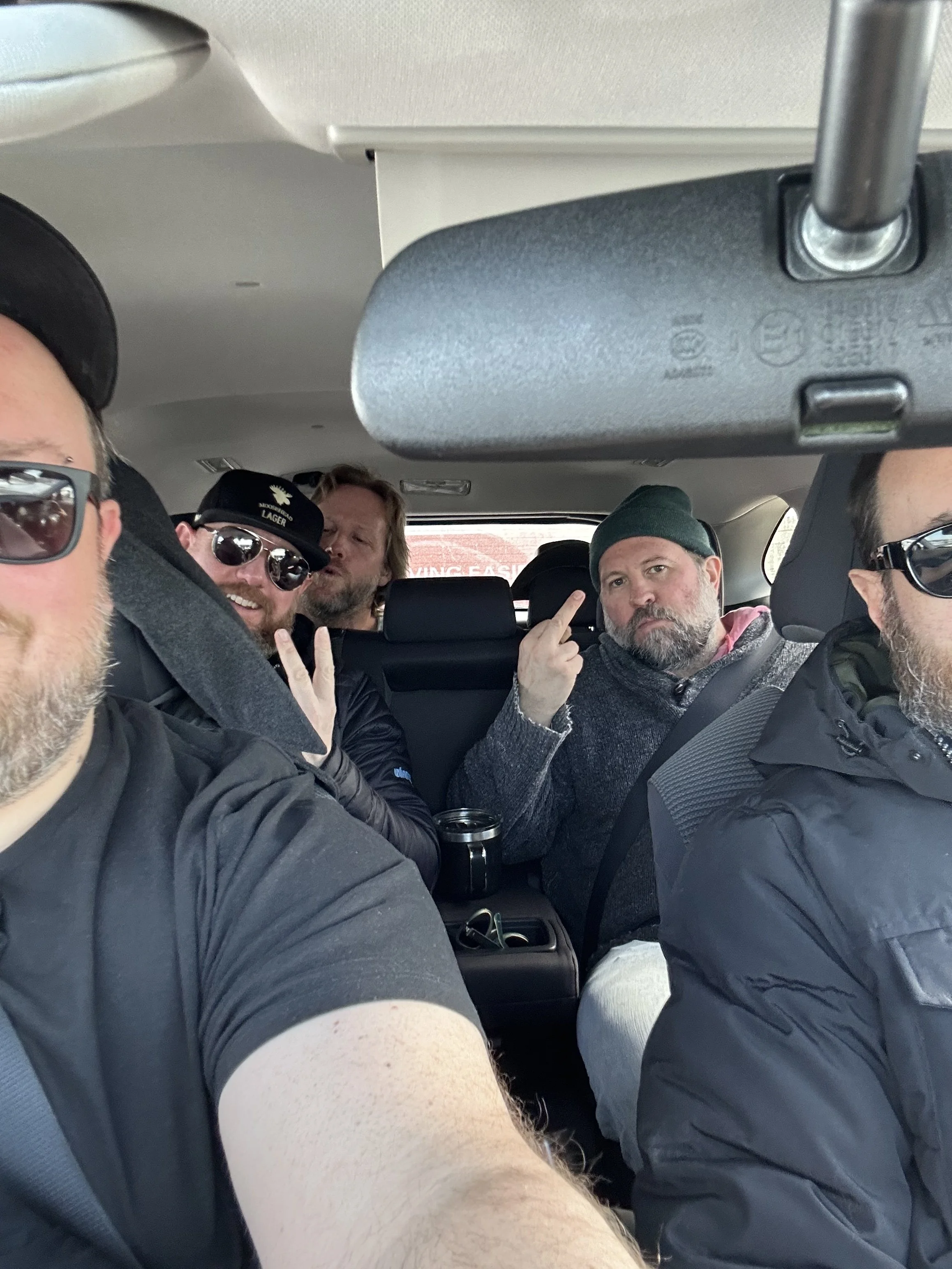 The band sitting inside a car, with some making gestures and wearing sunglasses, jackets, and hats.