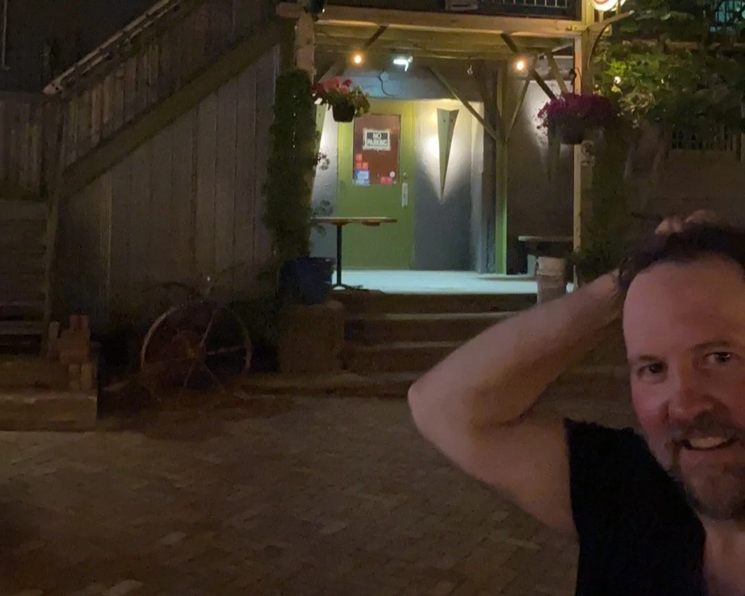 Nighttime outdoor scene with Dennis smiling in the foreground, and a wooden outdoor staircase leading to a brightly lit door adorned with signs, surrounded by plants and vintage decor.