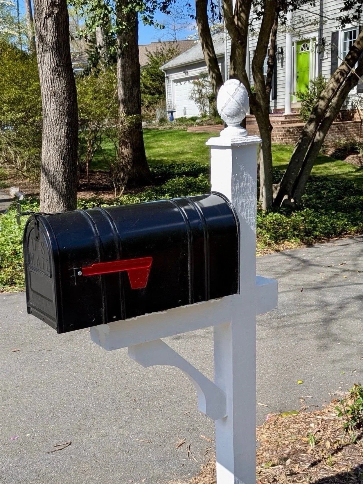 Black mailbox with red flag mounted on a white post along a suburban street, in front of trees and a house with a green door.