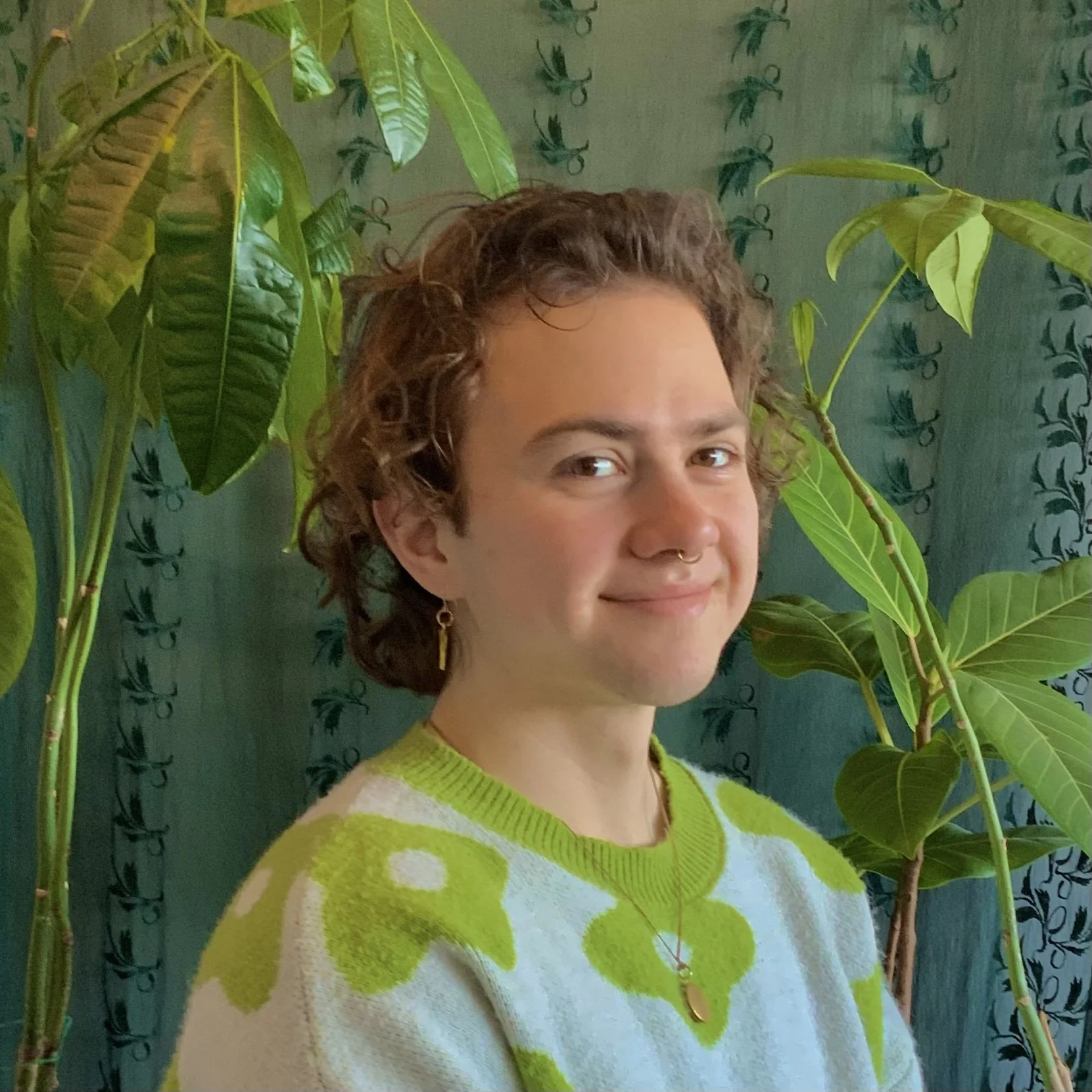 Portrait of the artist, Levi Angelo, smiling at the camera in front of his houseplants.  He has medium brown wavy hair and is wearing gold jewelry and a sweater with a green flower print.