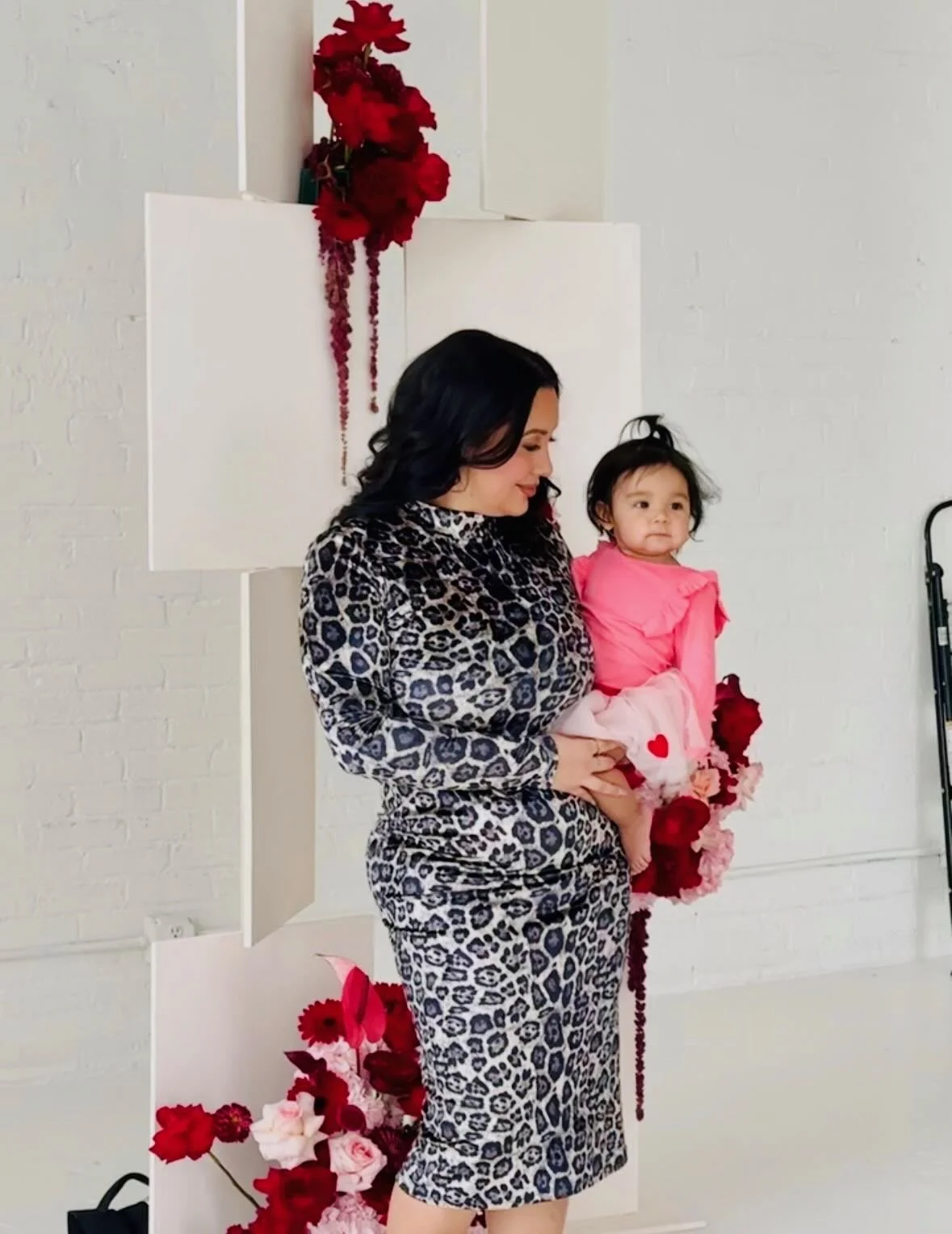 A woman holding a young girl in front of a white wall decorated with red and pink flowers.