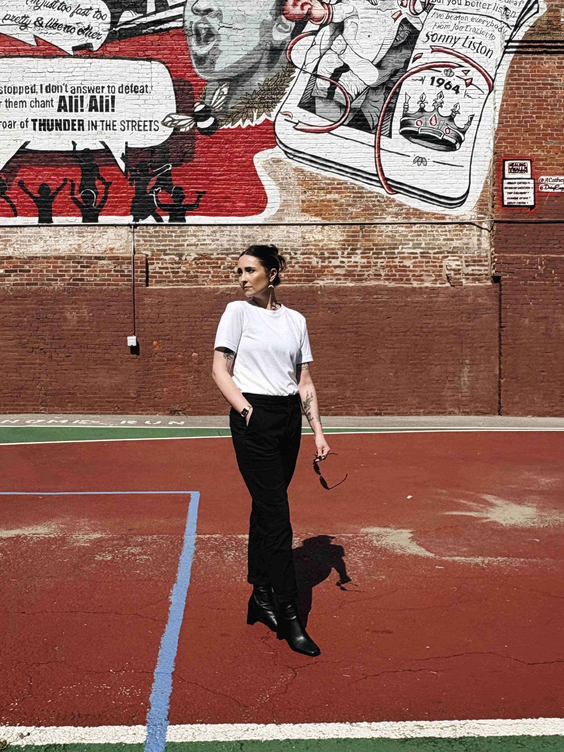 A woman standing on a red outdoor basketball court with a mural of a young boy, comic book elements, and a crown on a brick wall behind her. She is wearing a white T-shirt and black pants, holding sunglasses in her right hand, and has tattoos on her arms.
