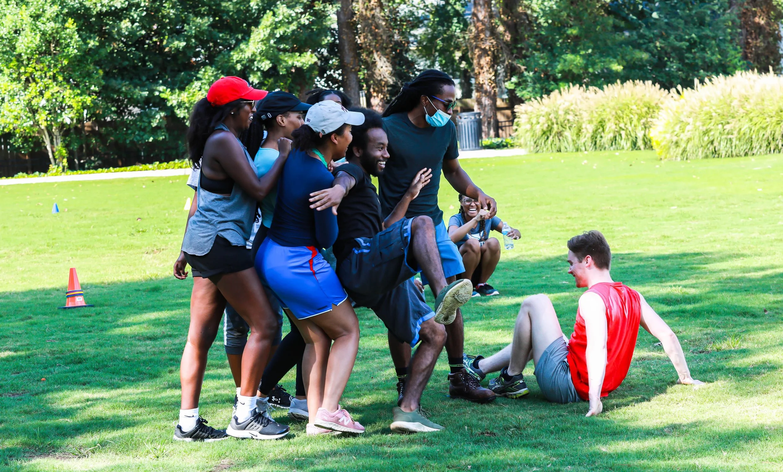 A group of people playing a game outdoors on a grassy field, some trying to lift a young man in a red sleeveless shirt who is sitting on the ground. Others are laughing and cheering, with trees and bushes in the background.