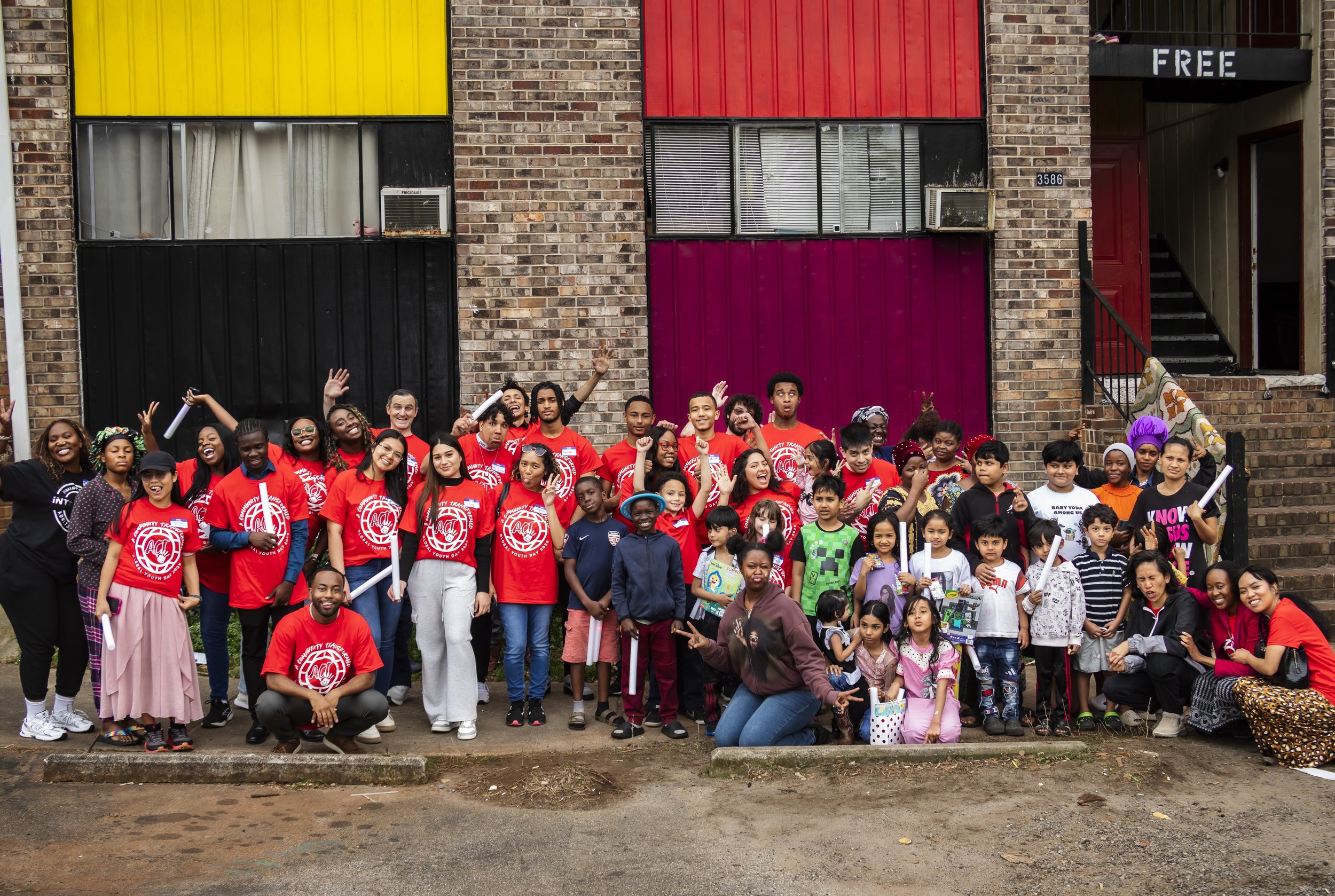 Group of diverse children and adults gathered outdoors in front of a brick building, smiling and waving, participating in a community event.