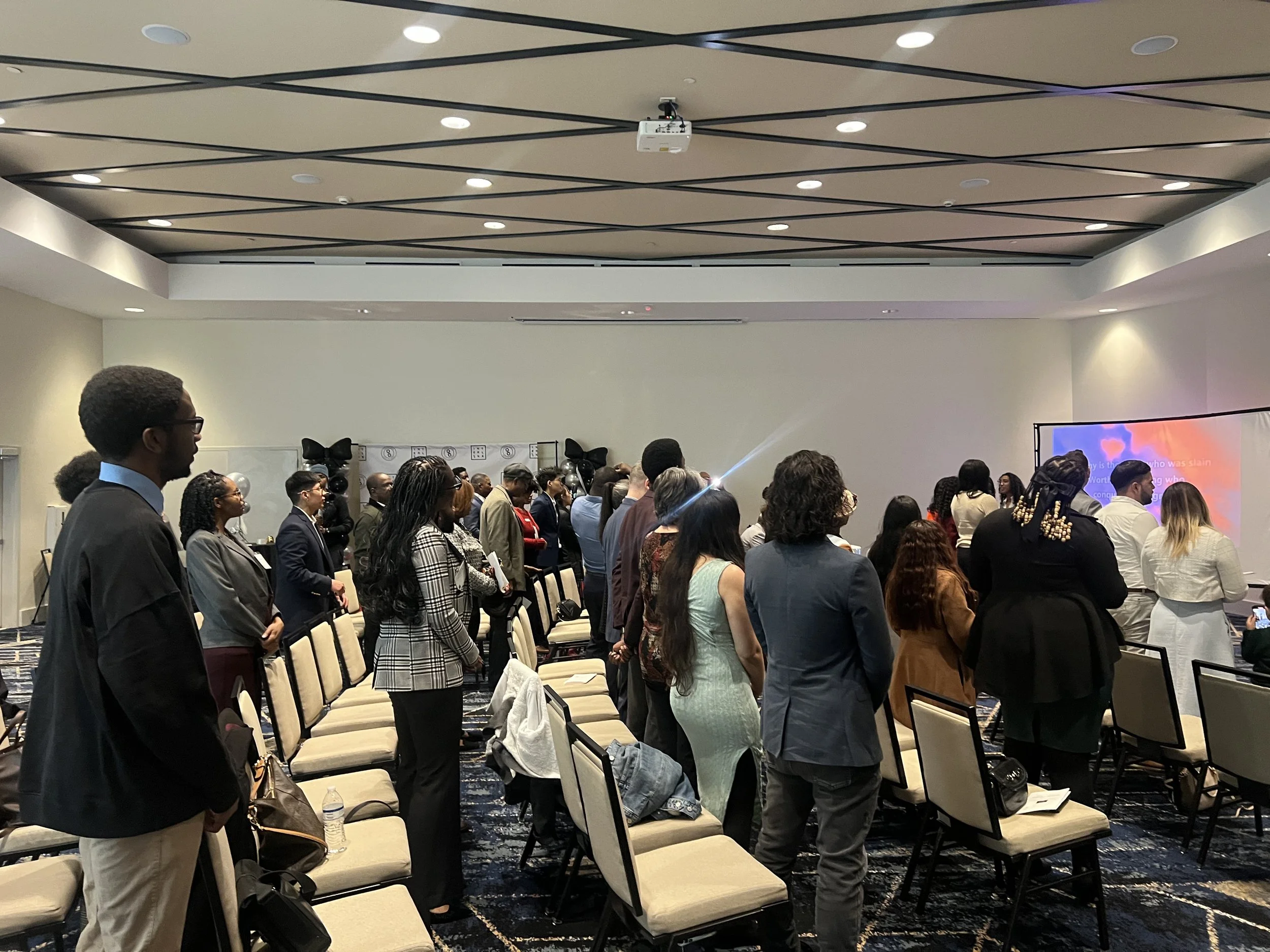 A large group of people standing and facing a stage in a conference room. The audience appears attentive, and the room is equipped with a ceiling-mounted projector and a large screen displaying colorful visuals.