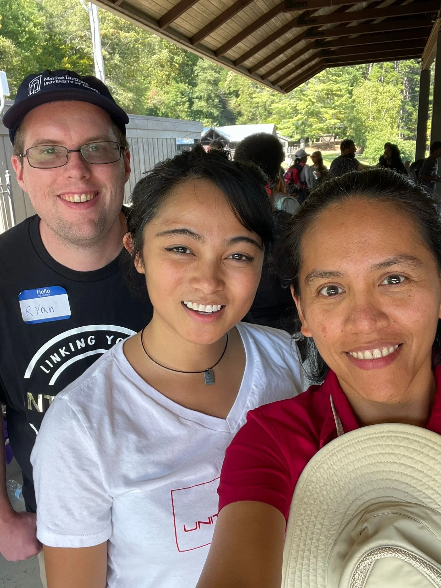 Three people standing outdoors under a wooden pavilion, smiling for a selfie. The background has many other people, trees, and tents or structures.