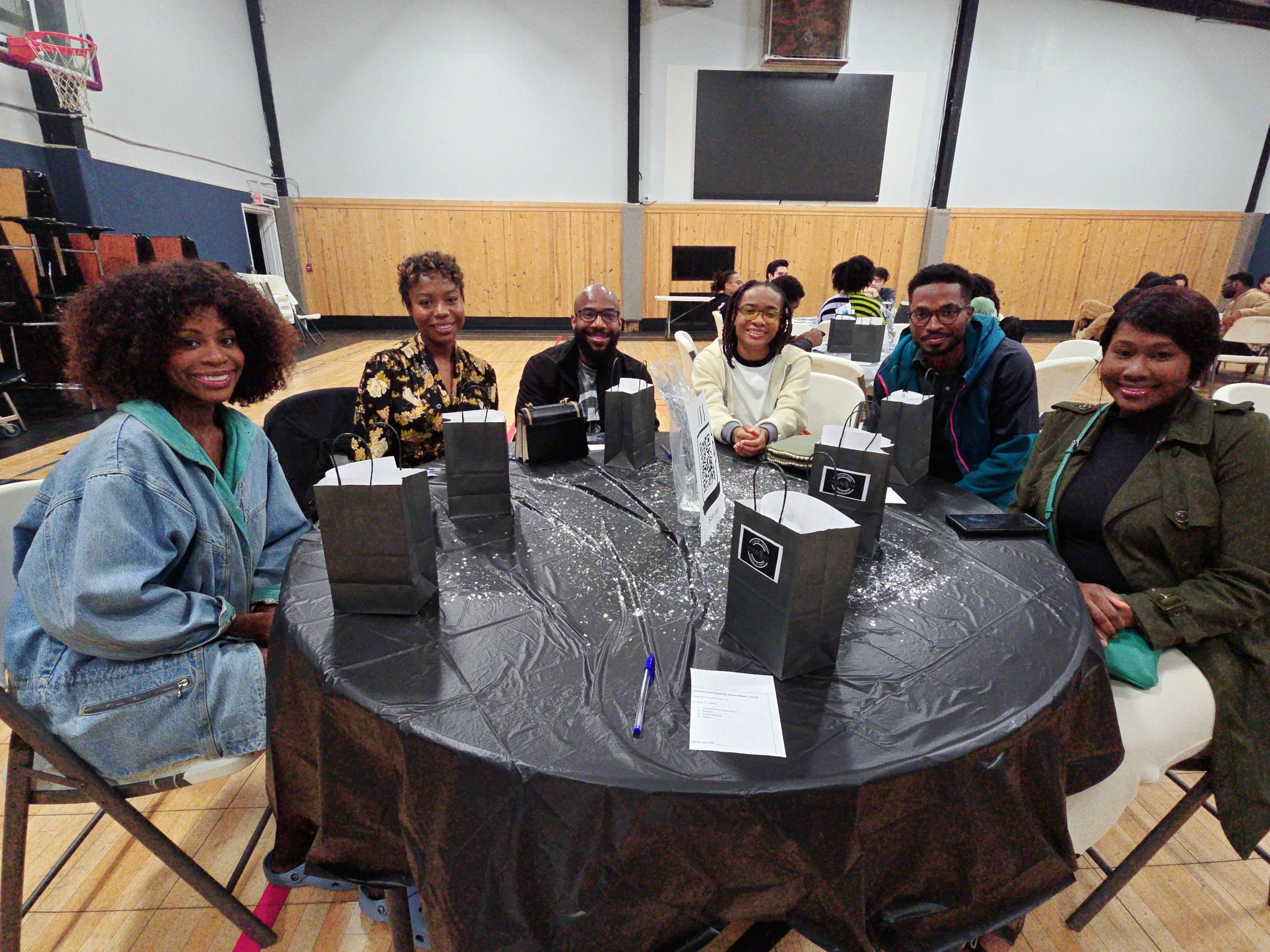 Six people sitting at a round table in a gymnasium, smiling at the camera. There are black gift bags on the table, and other tables and people in the background.