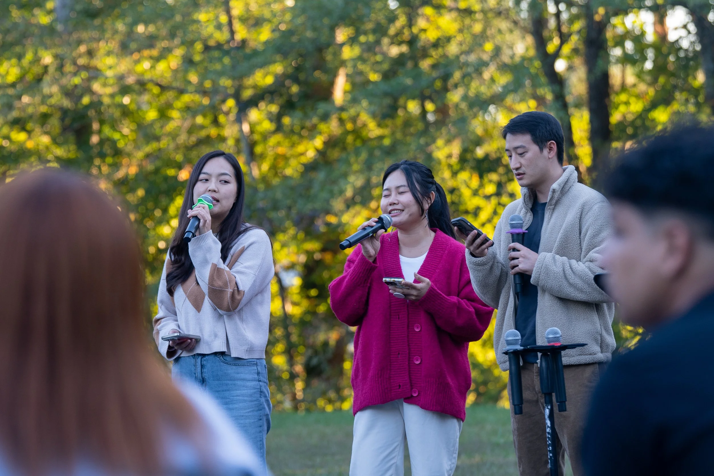 Three young adults standing outdoors with microphones and smartphones, surrounded by trees, participating in a speaking or singing event.