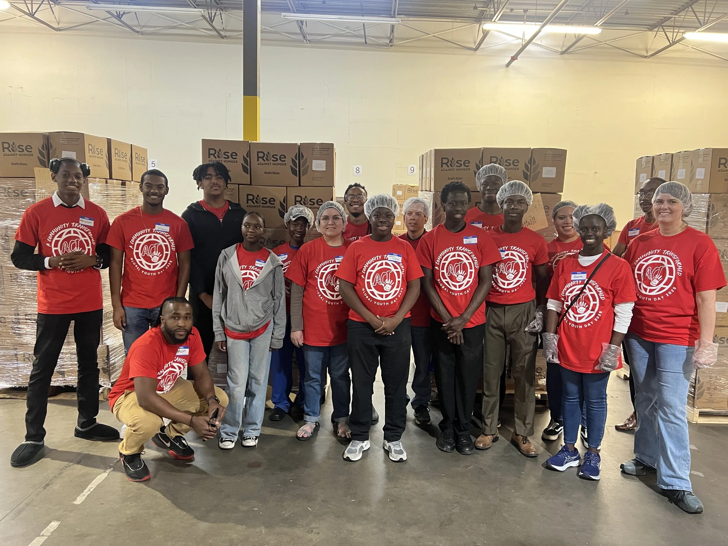 A group of people wearing red shirts with a logo and the words "Community Transformers Youth Day 2021" pose for a photo in a warehouse with stacks of boxes labeled "Rise Against Hunger." Some are wearing hairnets and gloves.