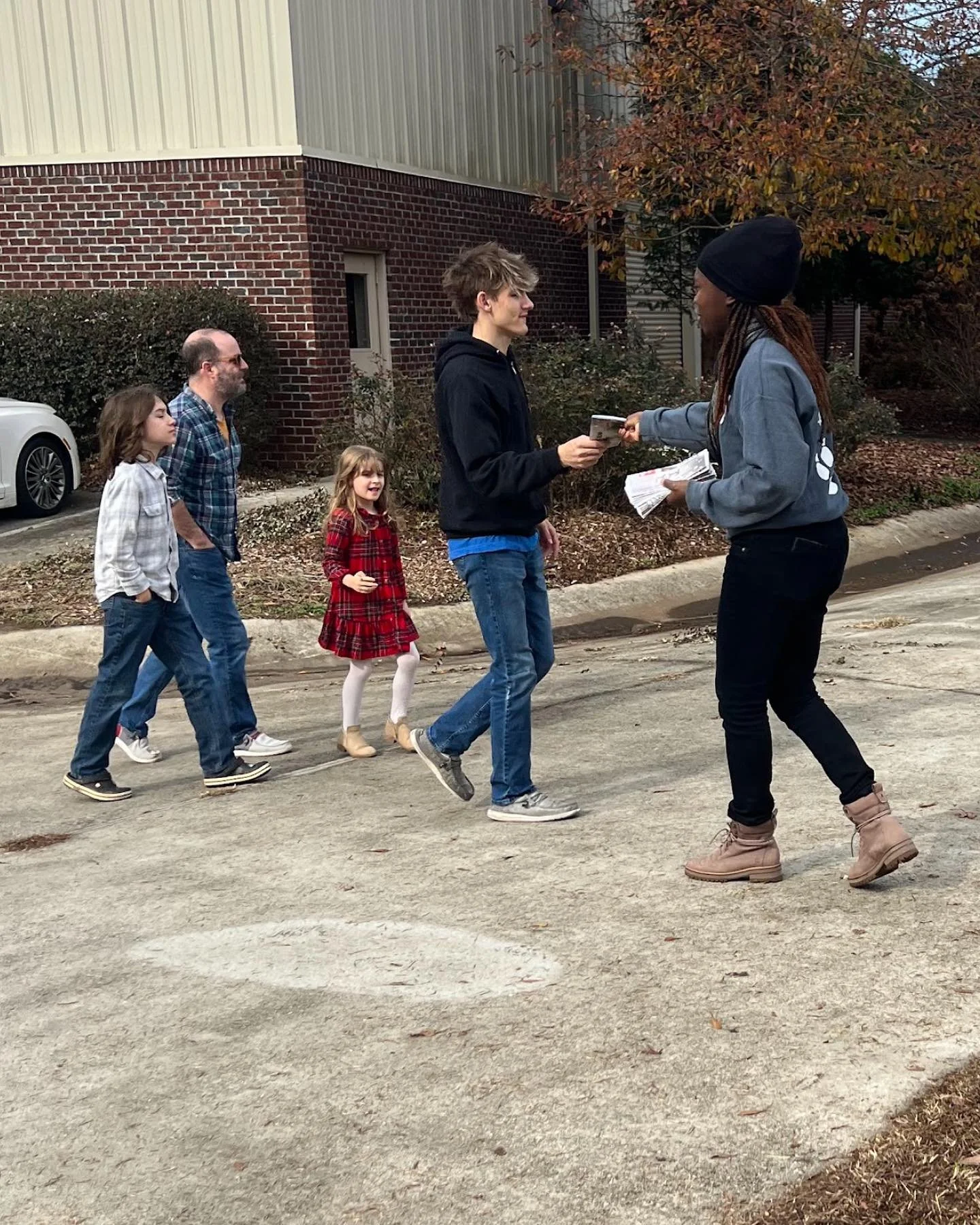 A group of people, including kids and adults, standing in line outdoors, with one person handing a flyer or pamphlet to a woman in a beanie. The setting appears to be outside a building with brick and metal siding, surrounded by fall foliage.