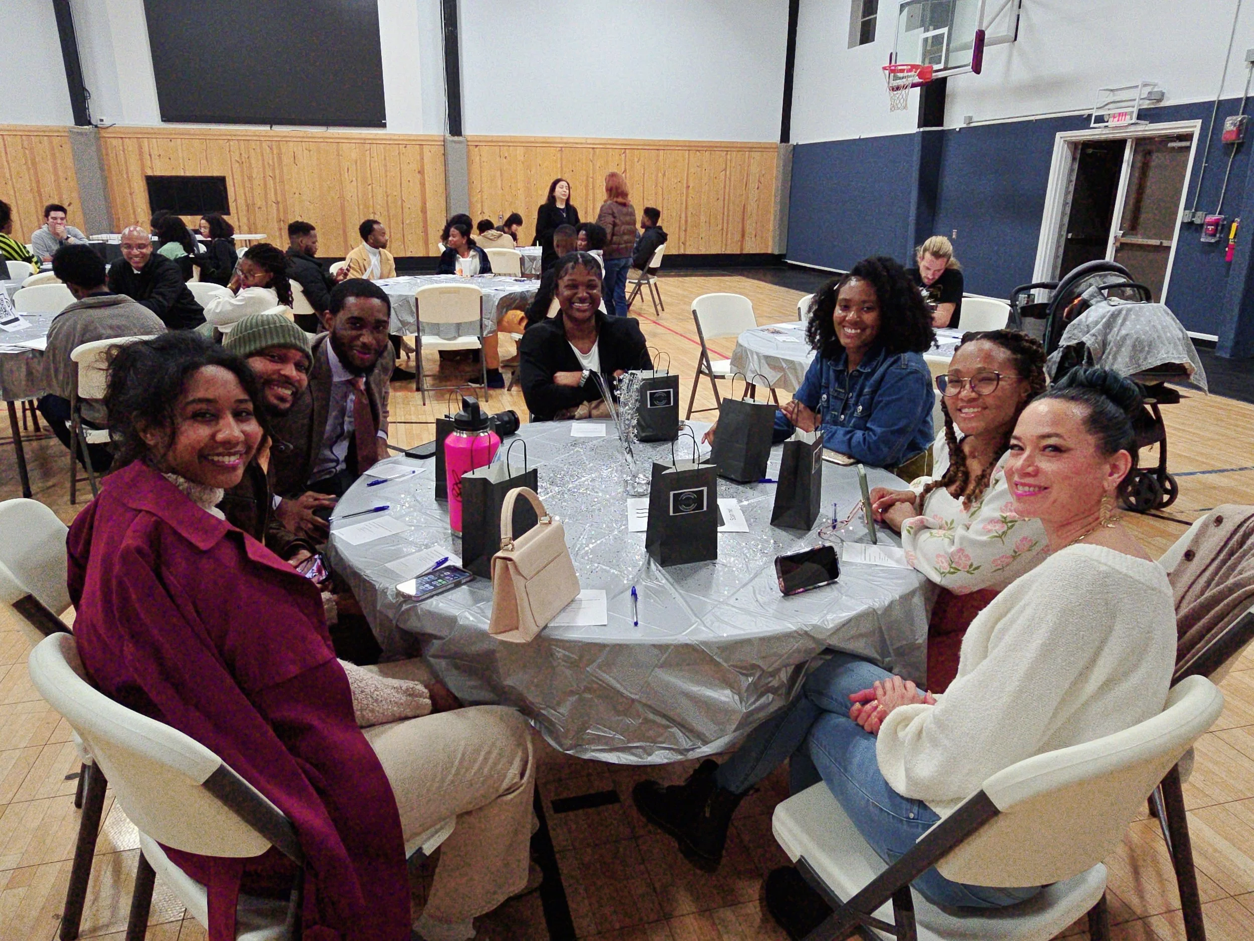 A diverse group of people gathered around a table at a social event in a gymnasium-like setting, smiling and engaging with each other.