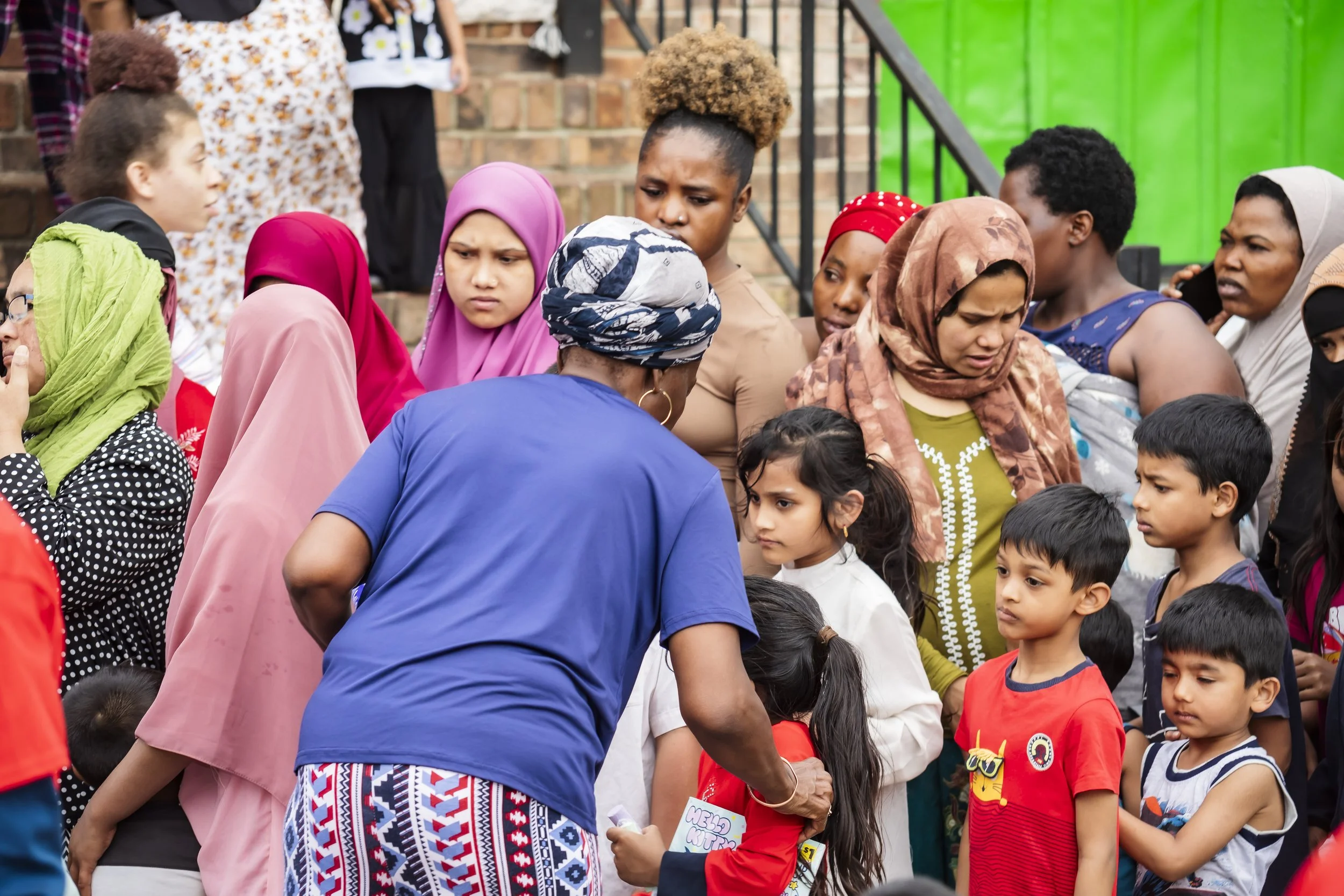 A diverse group of women and children gathered outdoors near brick steps, engaged in conversation or activity, with some wearing colorful headscarves and casual clothing.