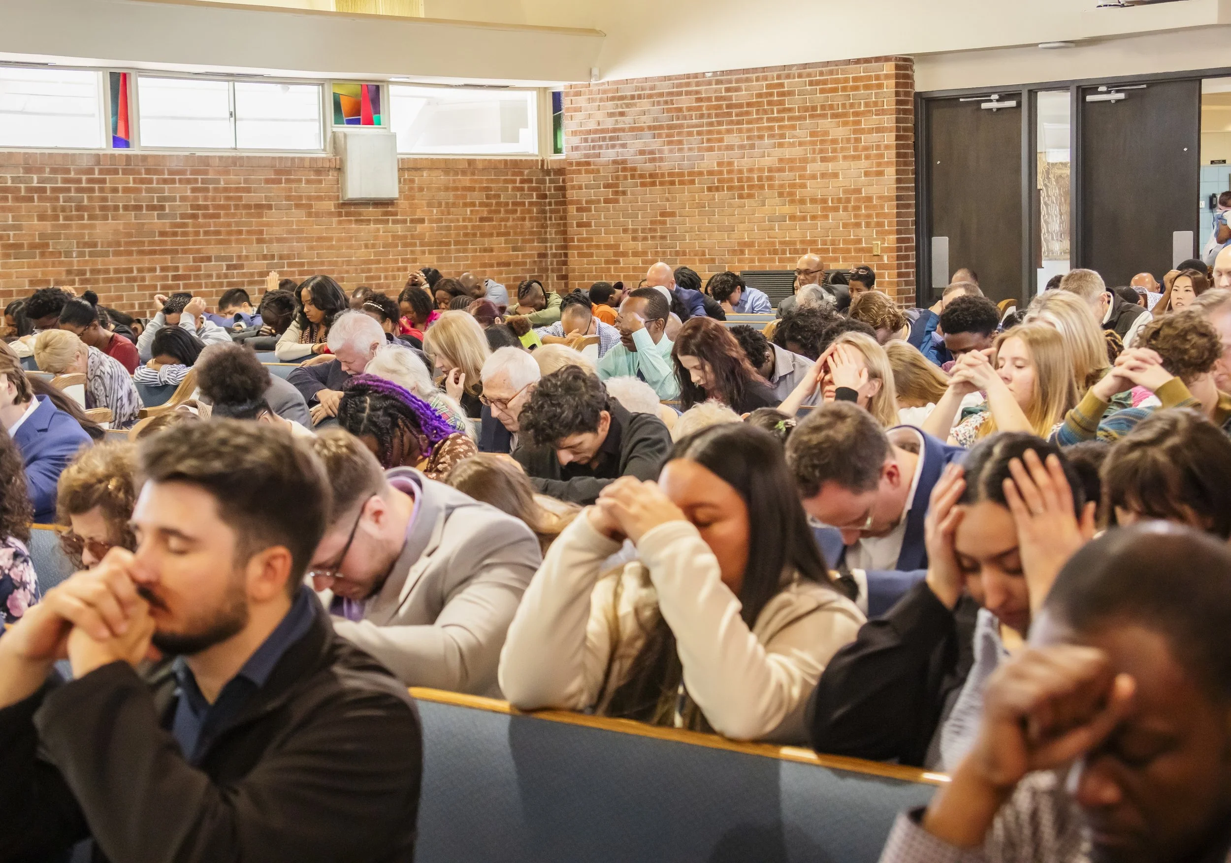 A large group of people gathered in a church or auditorium, holding their heads in prayer or reflection, with brick walls and stained glass windows in the background.
