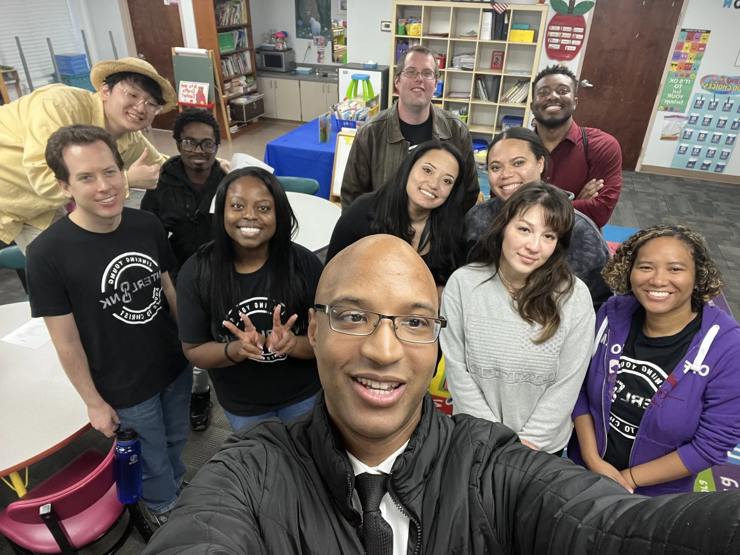 A group of people taking a selfie in a colorful classroom or library setting with bookshelves, posters, and tables in the background.