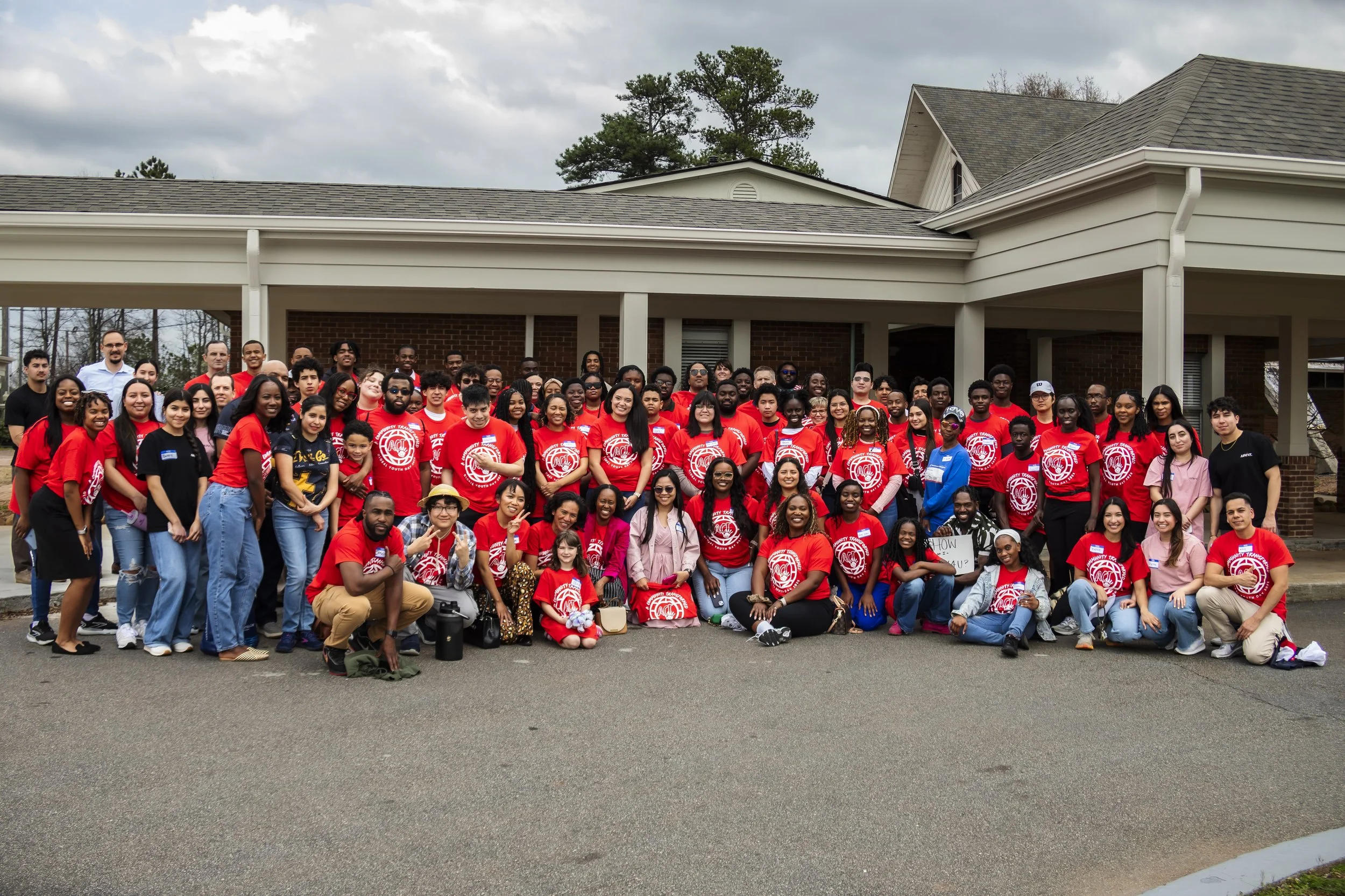 Group of diverse people, many wearing red shirts, gathered outside in front of a building for a group photo on a cloudy day.