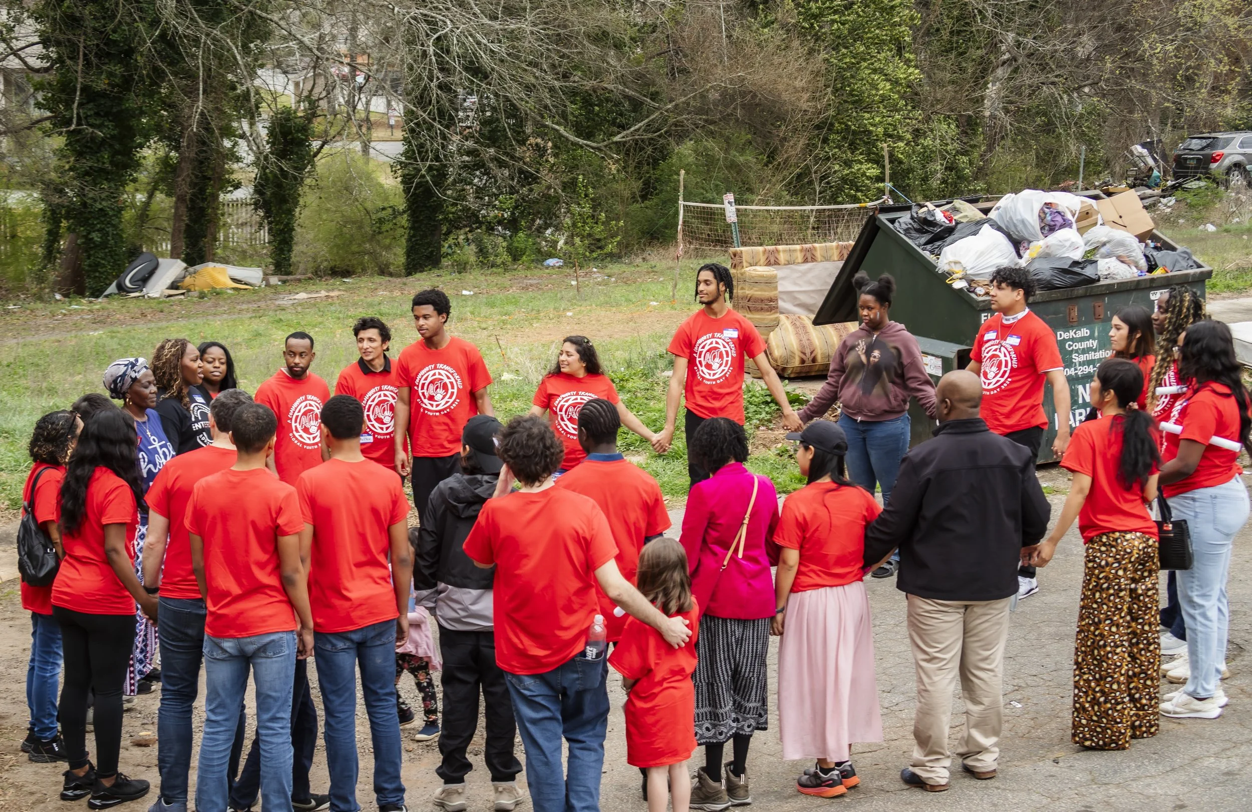 A diverse group of people, some wearing red shirts, holding hands and standing in a circle outdoors during daytime, with a large dumpster overflowing with trash in the background, indicating a community cleanup event.