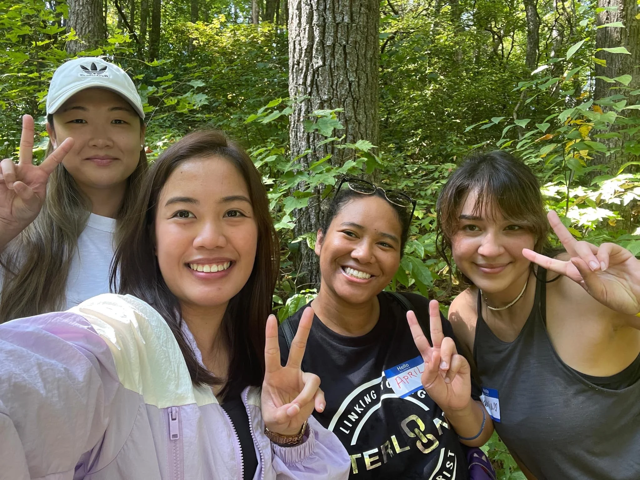 Four women taking a selfie outdoors in a wooded area, smiling and making peace signs.