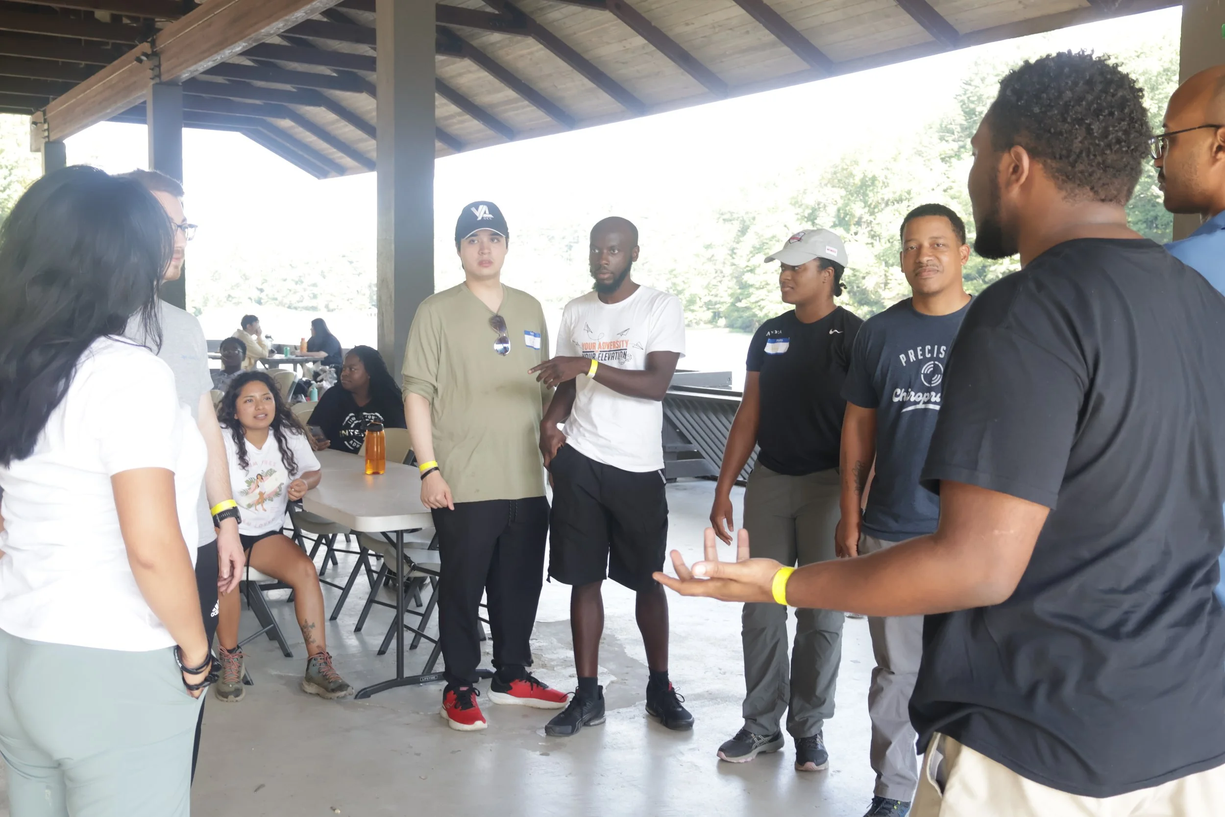 Group of diverse people engaged in a discussion or activity outdoors under a pavilion, with some seated at tables and others standing.