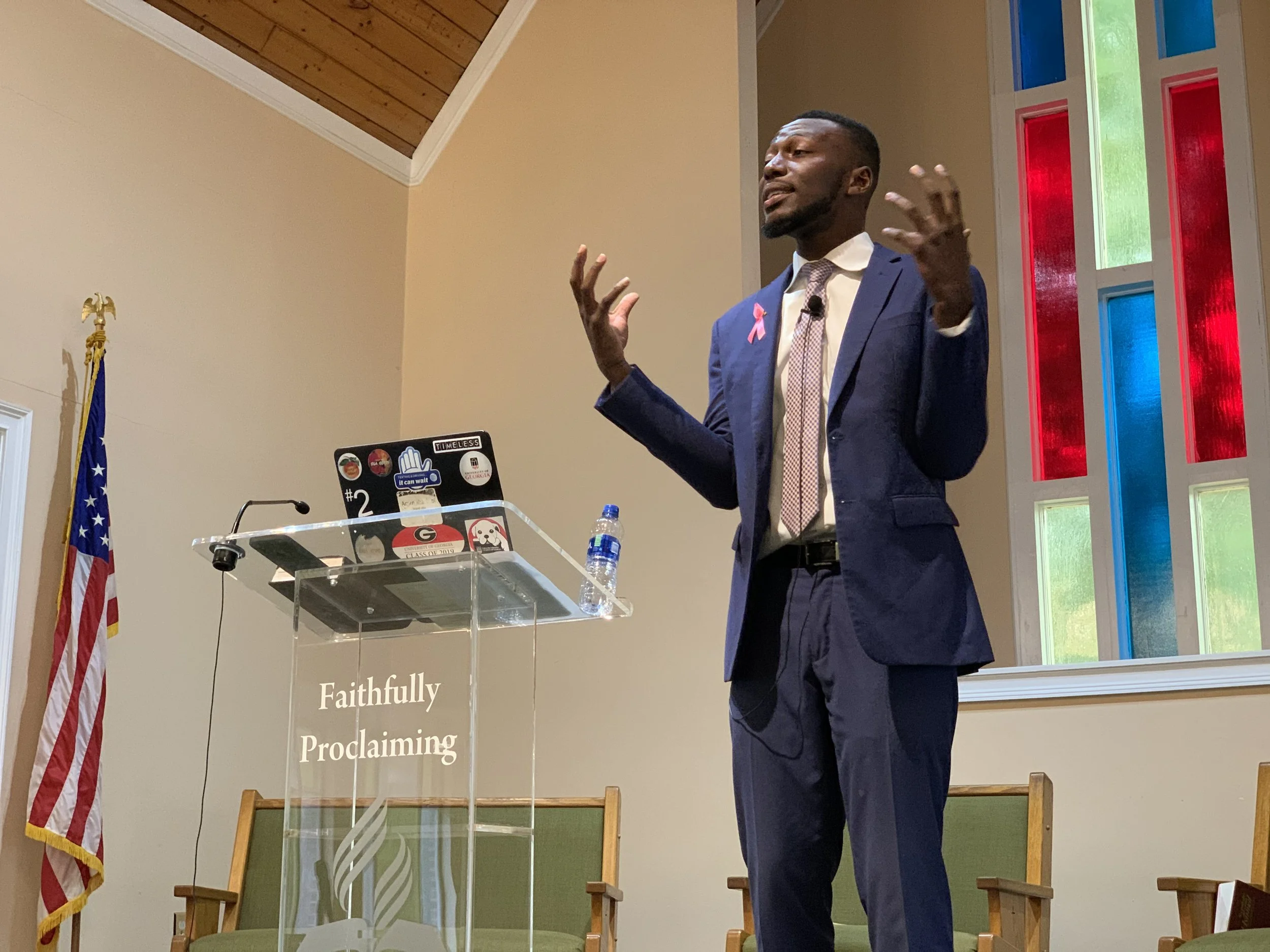 A man in a blue suit and white shirt is giving a presentation at a church or religious event, with colorful stained glass windows behind him and an American flag to his left. He is gesturing with his hands, and there is a clear podium with various stickers on it in front of him.