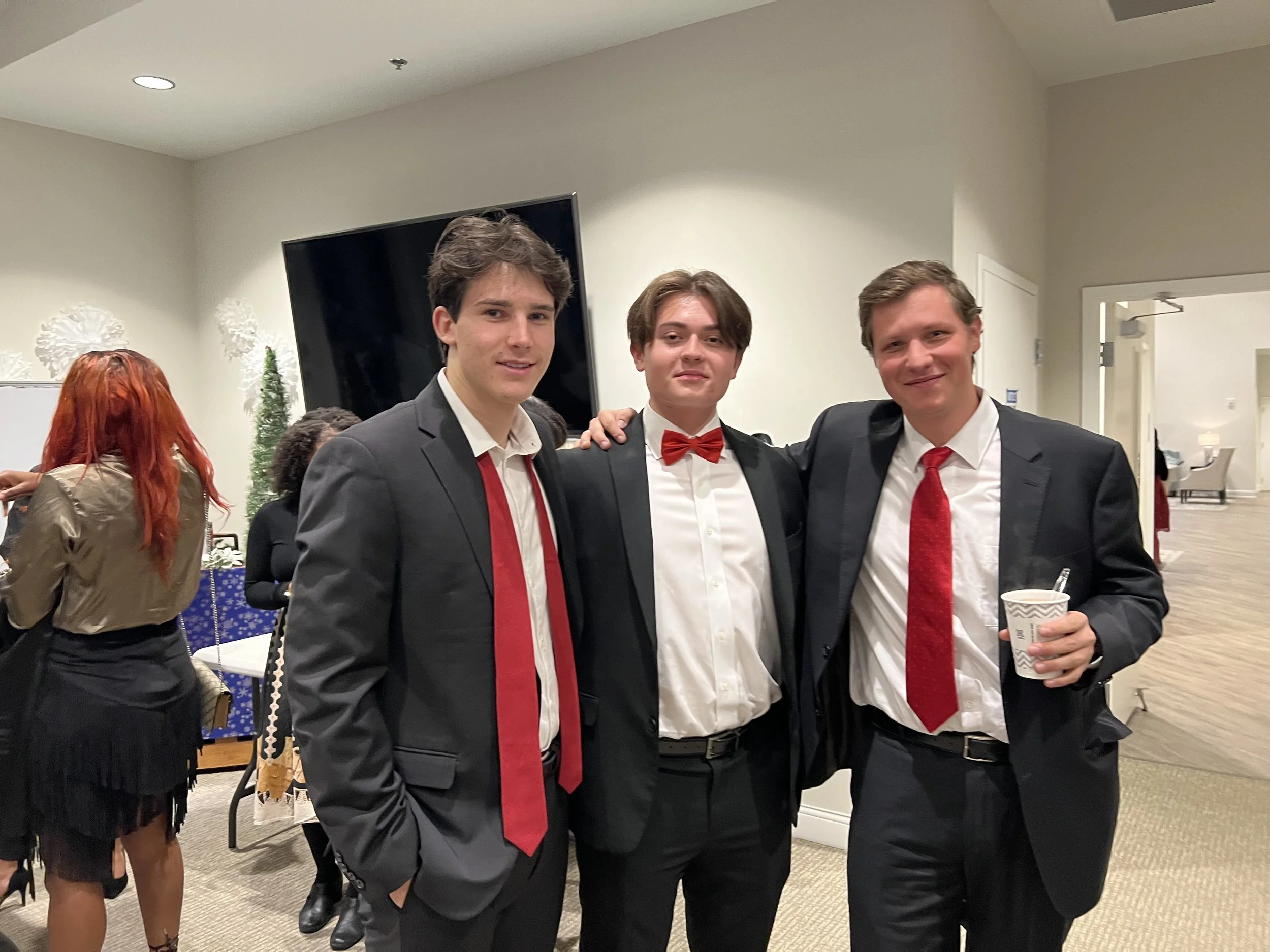Three young men in formal attire posing together at a social event, with one holding a cup.