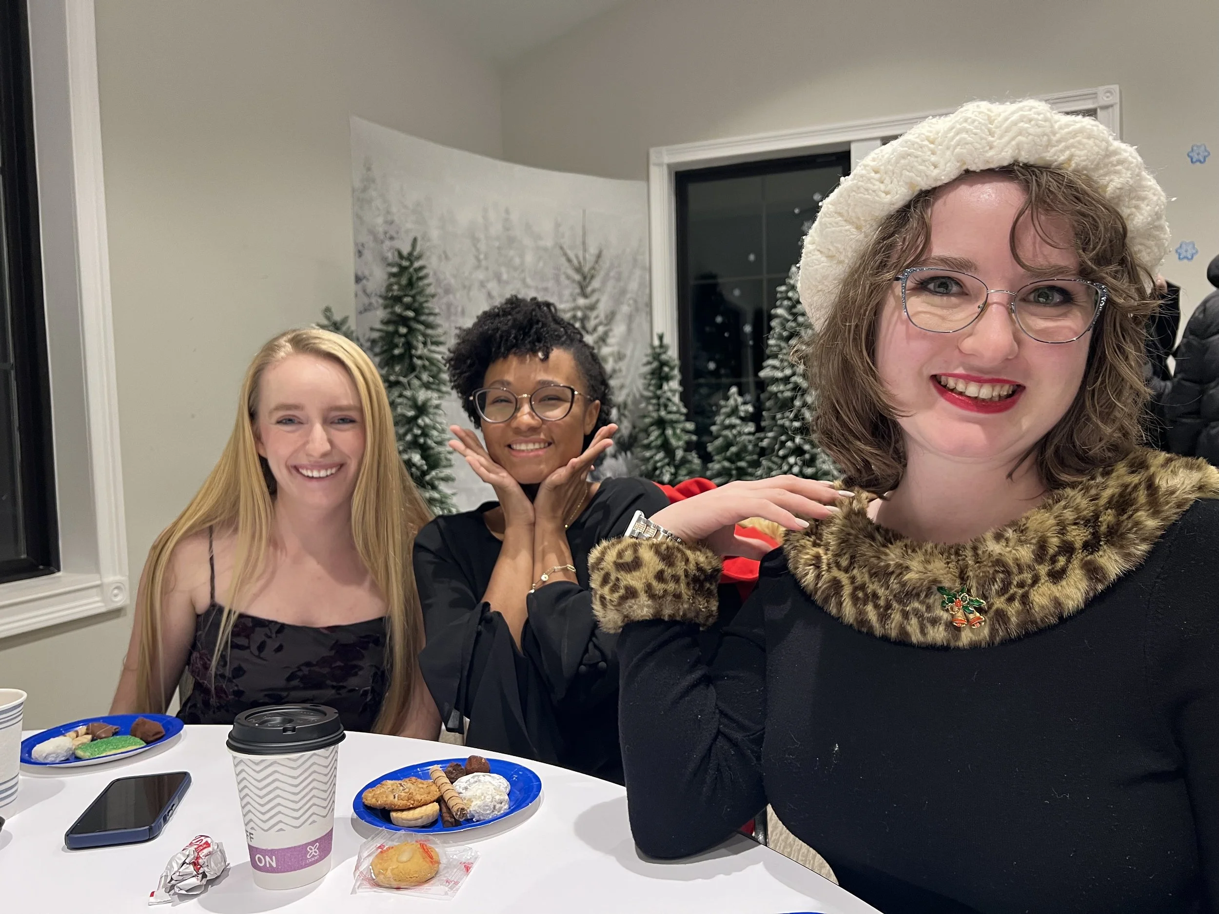 Three women smiling at a holiday gathering, with a snowy forest scene with evergreen trees in the background. One woman in the foreground wears glasses and a fuzzy hat, while the other two women behind her have long hair and are also smiling.