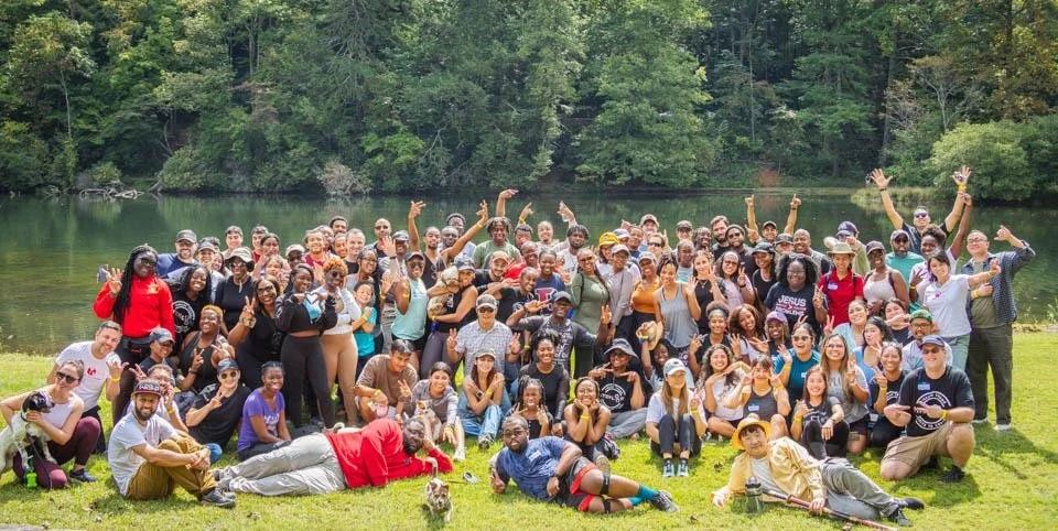 A large diverse group of people gathered outdoors near a lake with trees in the background, posing for a group photo on a sunny day.