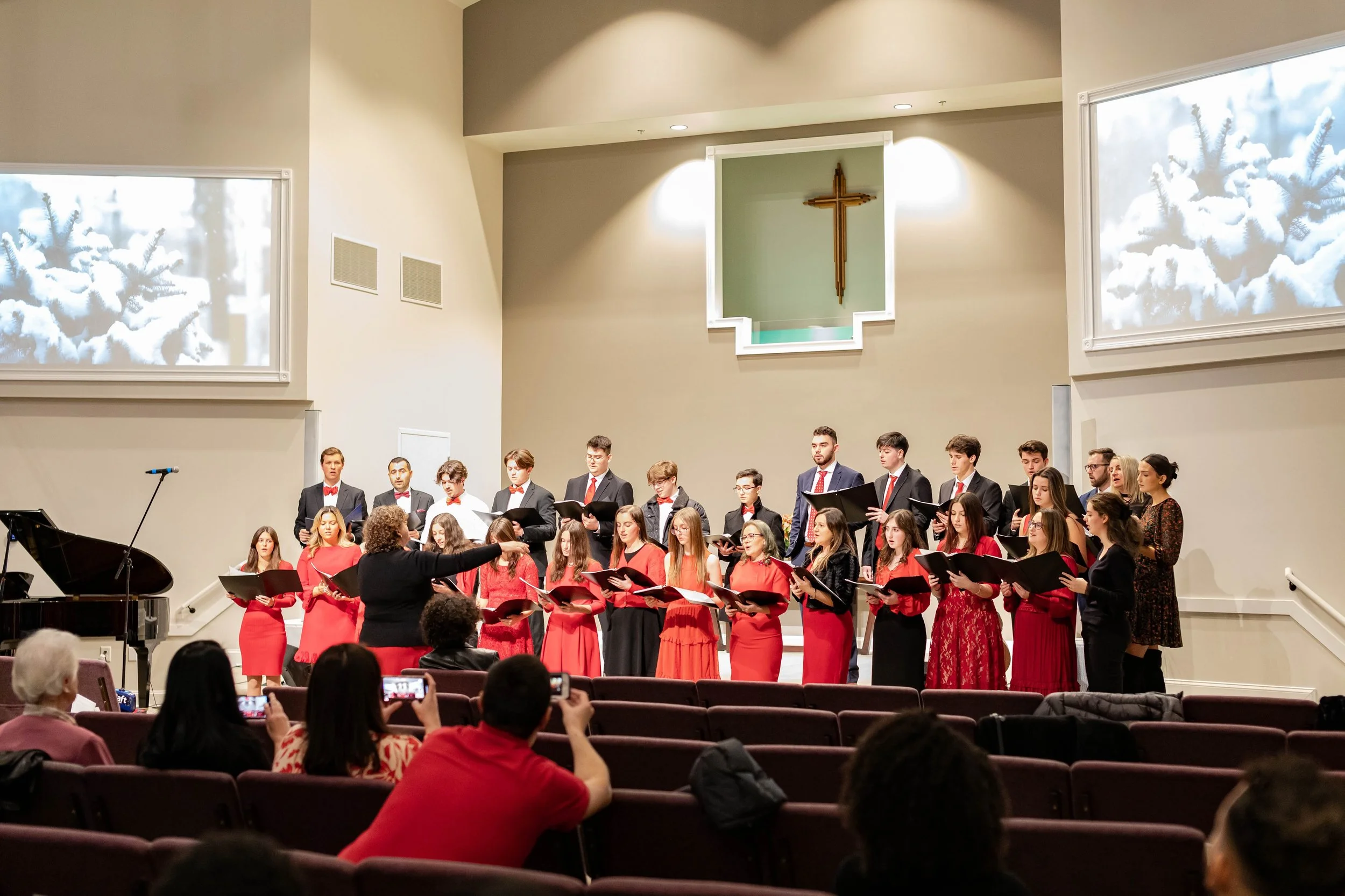 A choir performing in a church or concert hall with an audience taking photos, a conductor leading, and a large cross on the wall behind them.